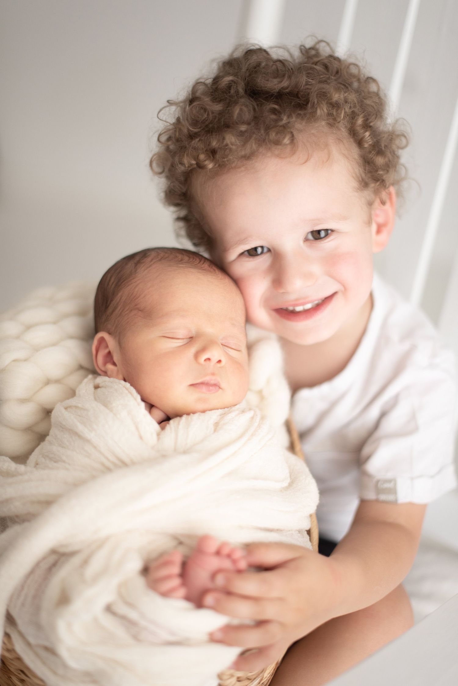 A newborn baby swaddled in white being held by a sibling with curly hair against a light background.
