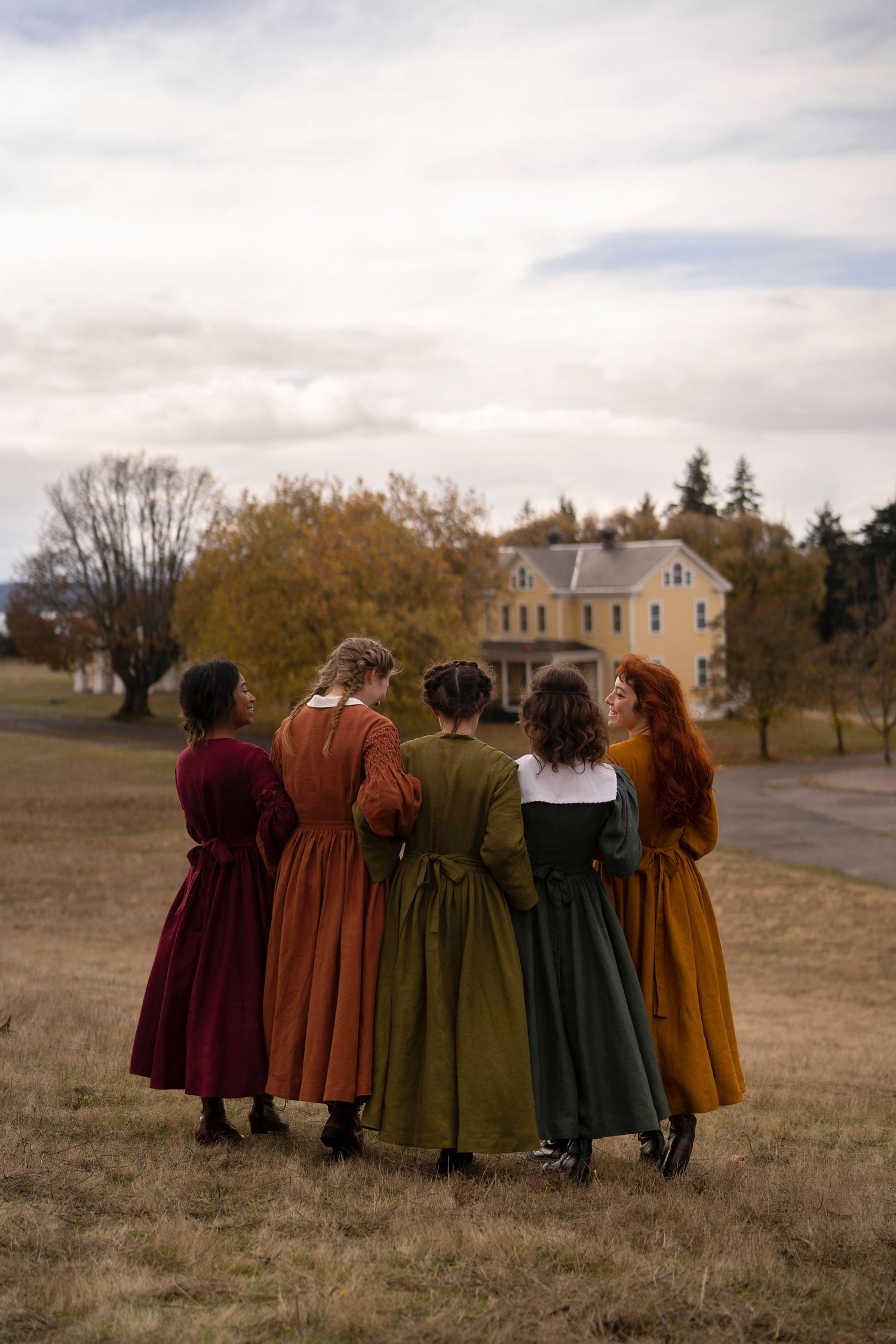 A group in colorful period dresses stands with backs turned, facing a pastoral scene with a yellow house.