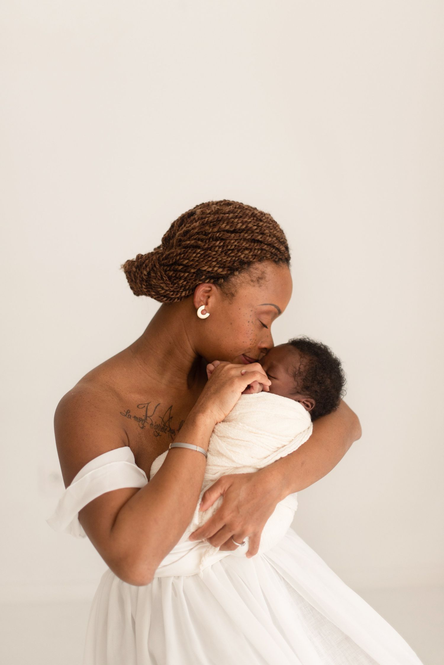 Tender moment between mother and baby in white dress against light background.