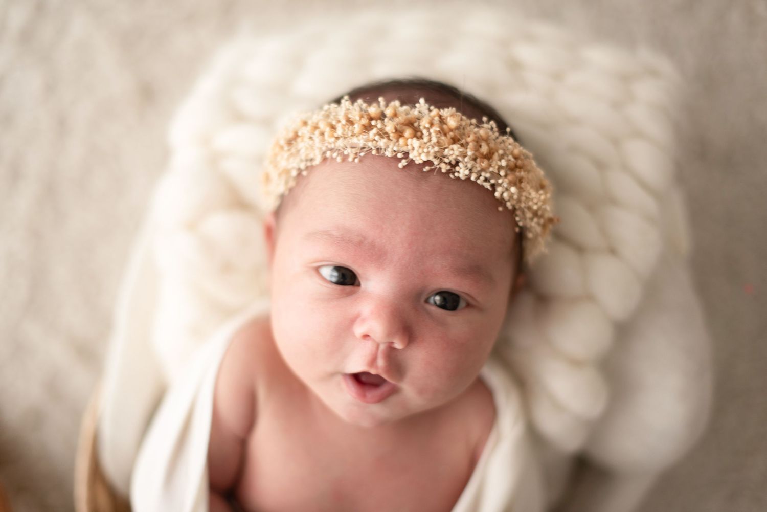 Baby wearing delicate golden headband lying on soft white blanket during newborn photography session.