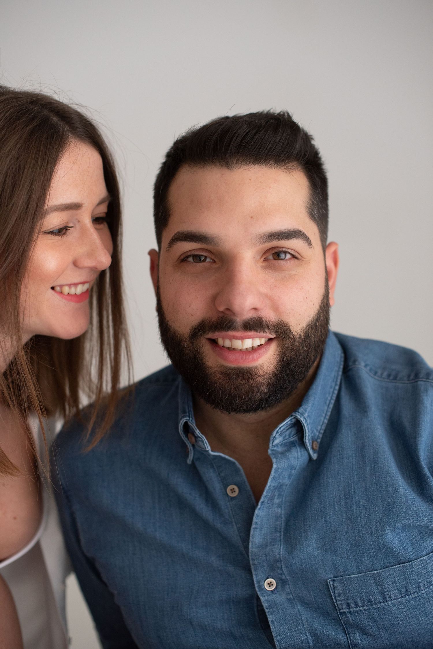 A couple smiles warmly at the camera while posing together against a neutral background.