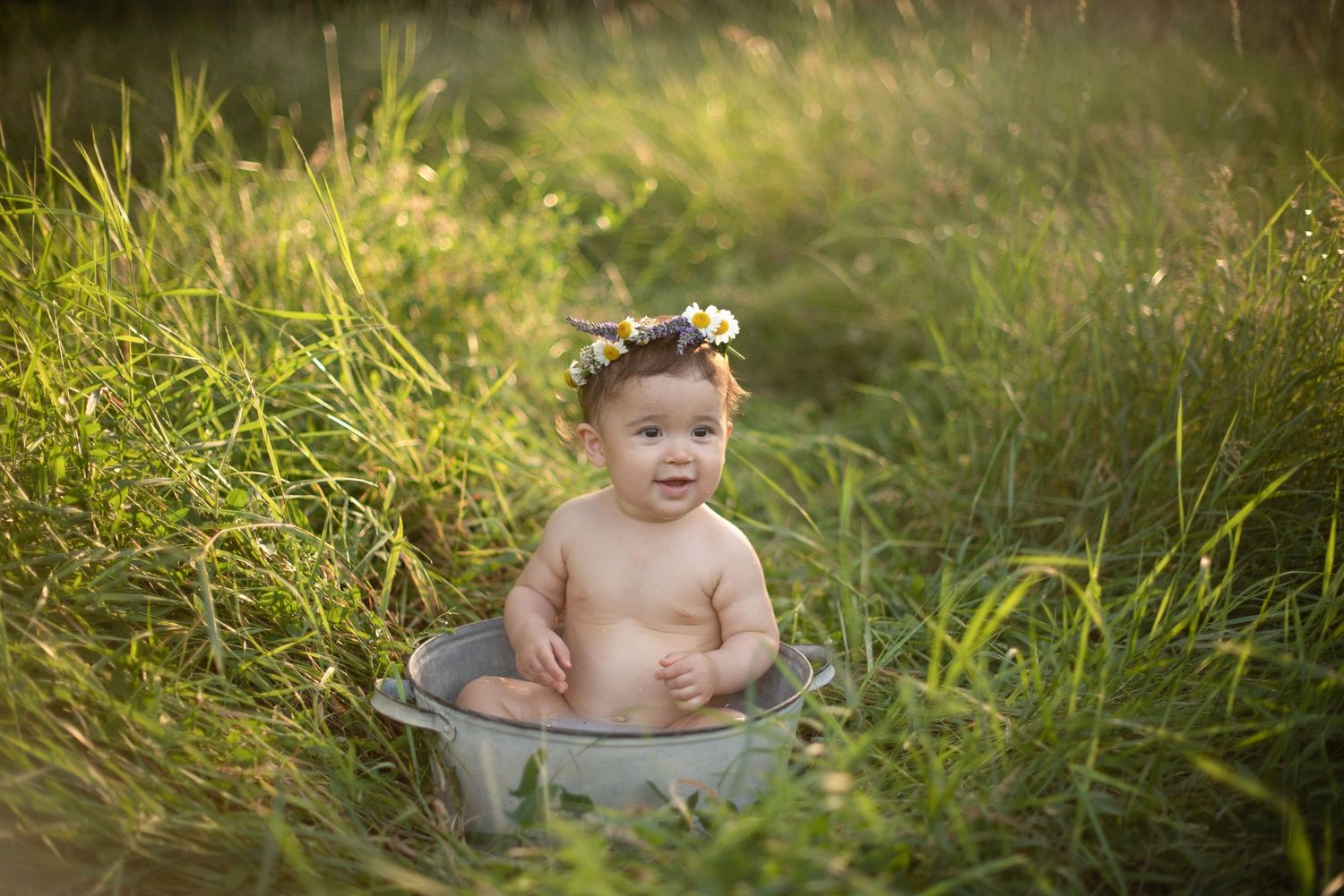 Baby wearing flower crown sits in metal basin among tall grass during sunset.