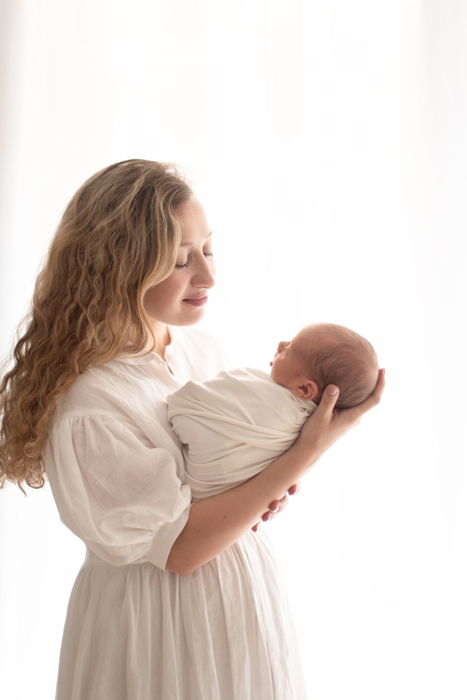 Woman in white dress holding swaddled newborn in soft backlit portrait.