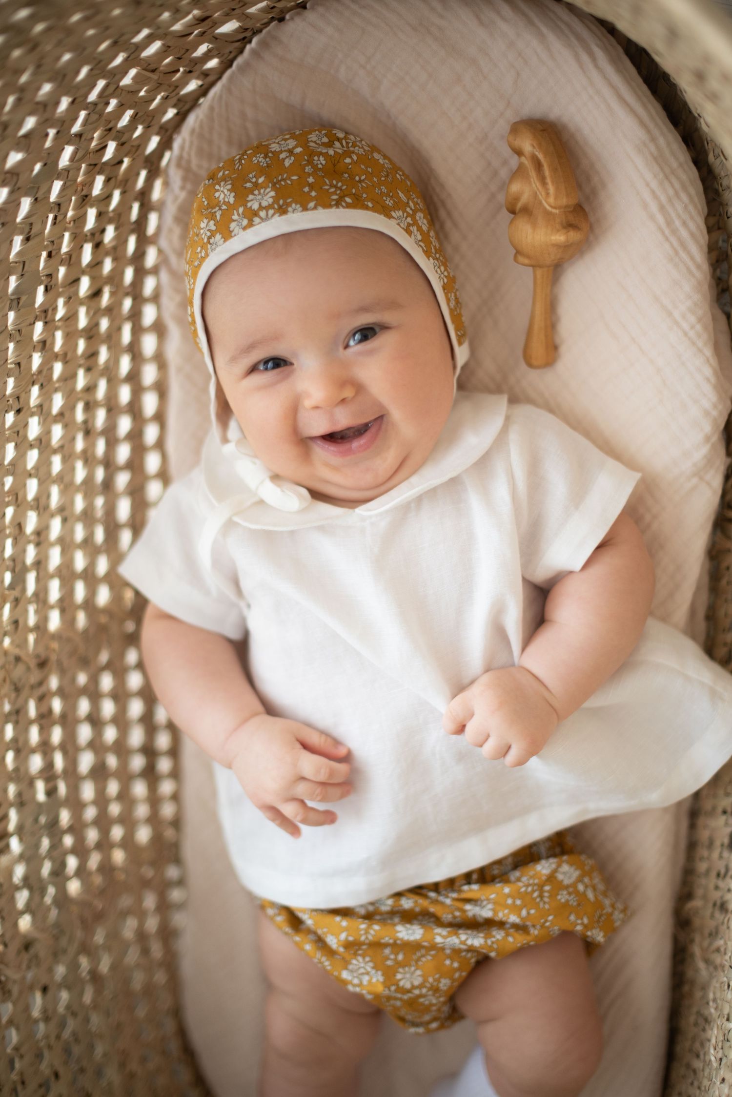 Baby wearing white top and yellow floral bonnet lying in woven basket with wooden toy.
