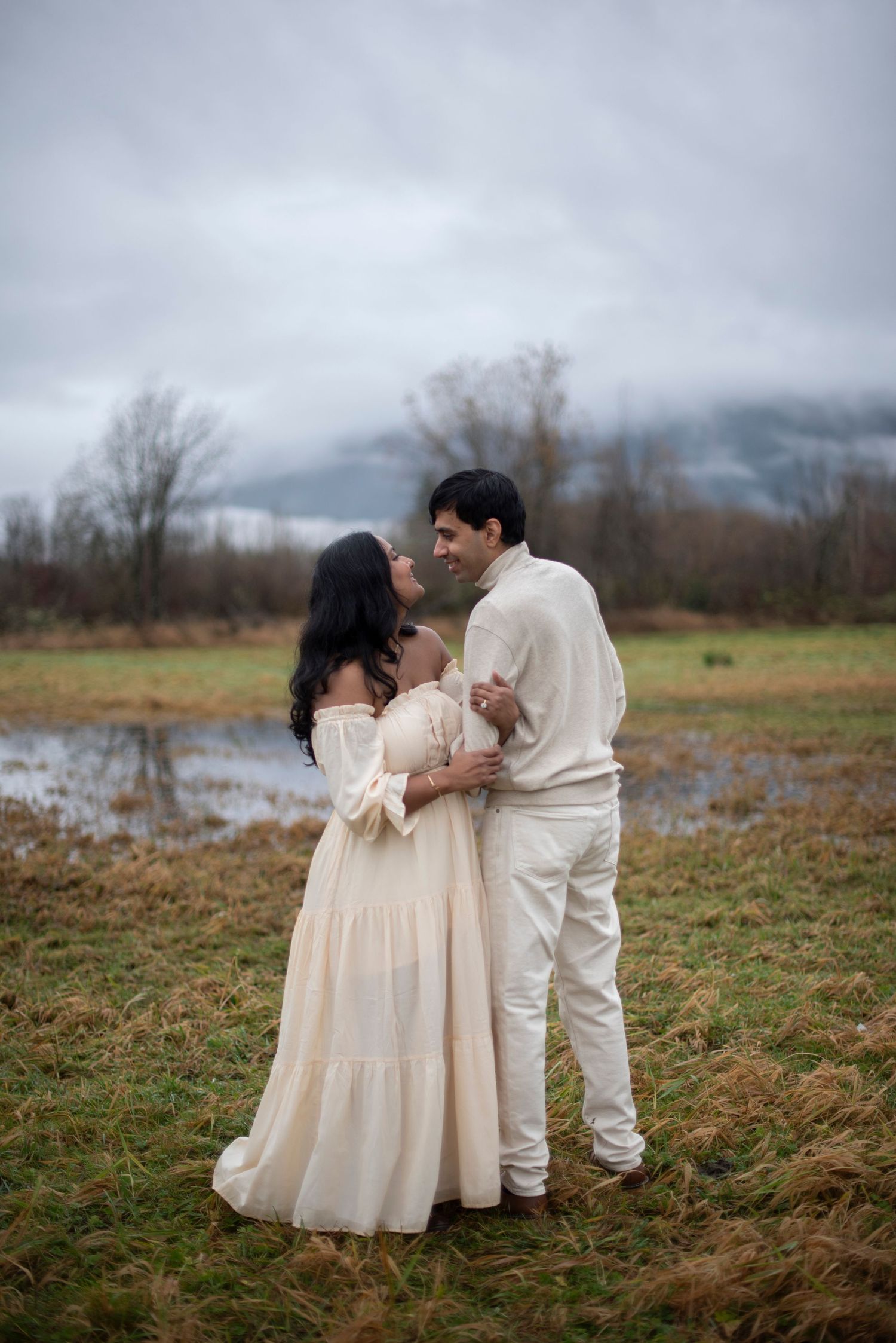 A couple in cream-colored outfits share an intimate moment in a misty field during an outdoor photoshoot.