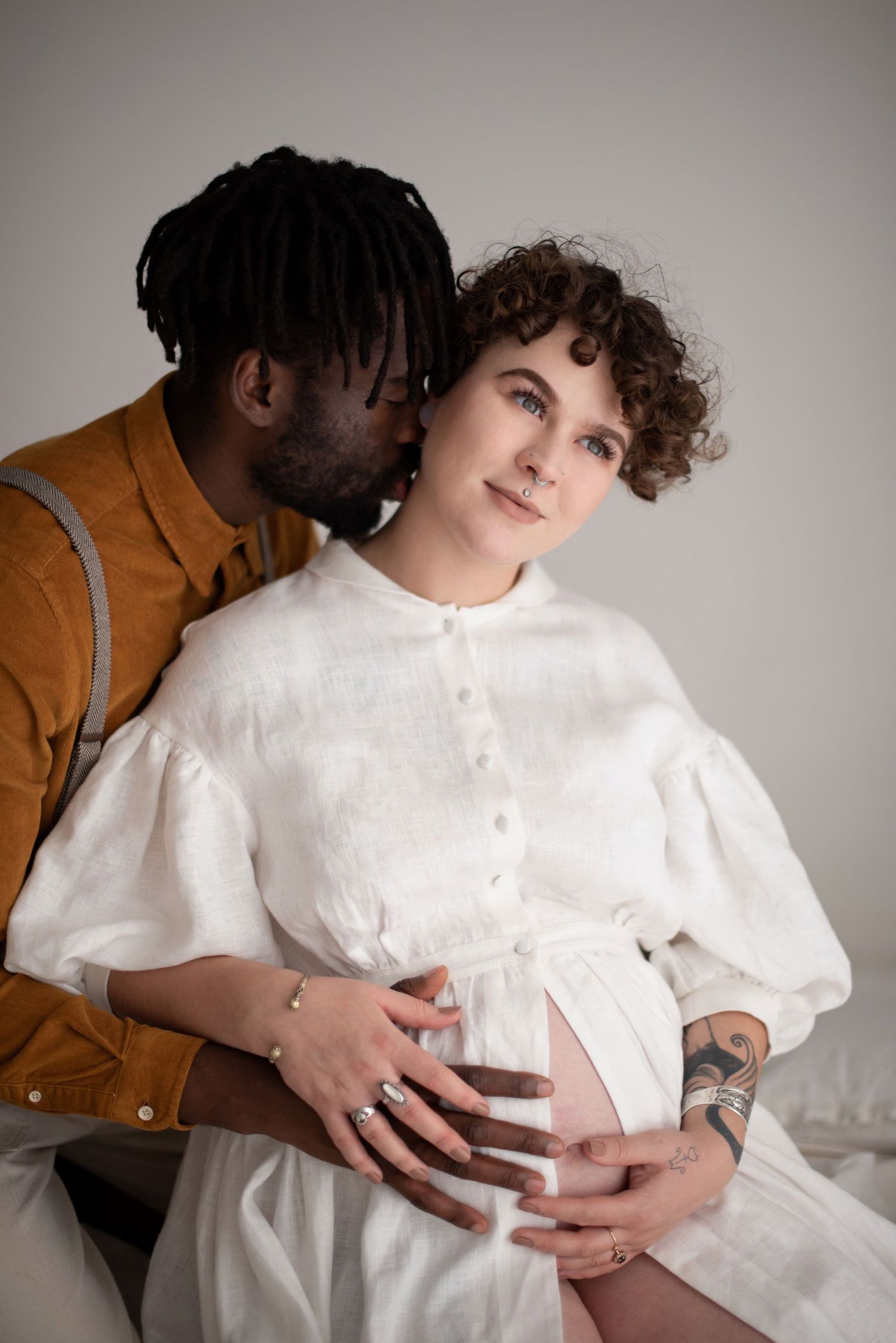 Intimate maternity portrait with couple embracing, person in white dress showing baby bump while partner whispers from behind.