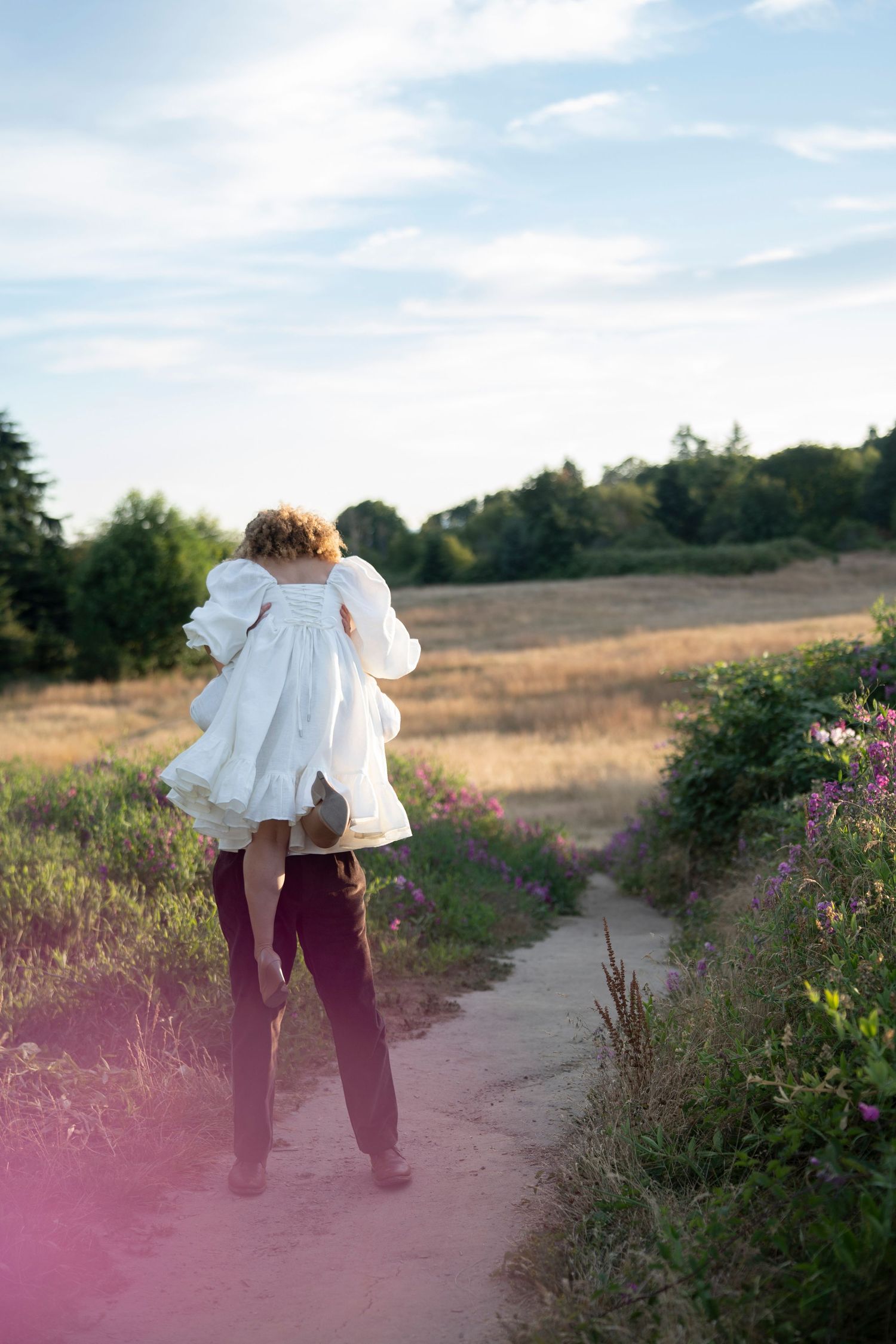 Figure walking on a dirt path through a sunny meadow at golden hour with pink lens flare effect in the foreground.