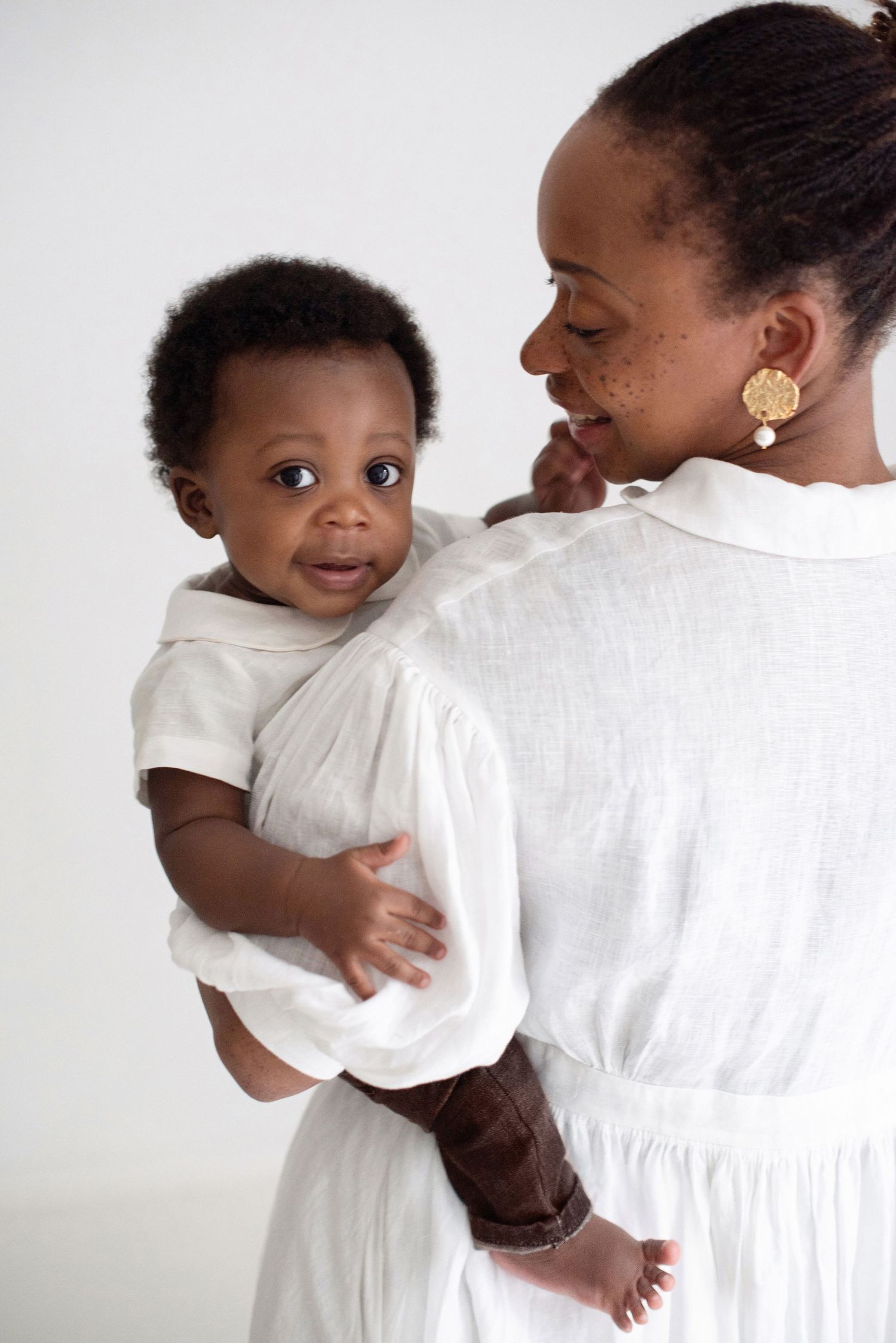 A tender moment between parent wearing white dress and small child in white shirt against light background.