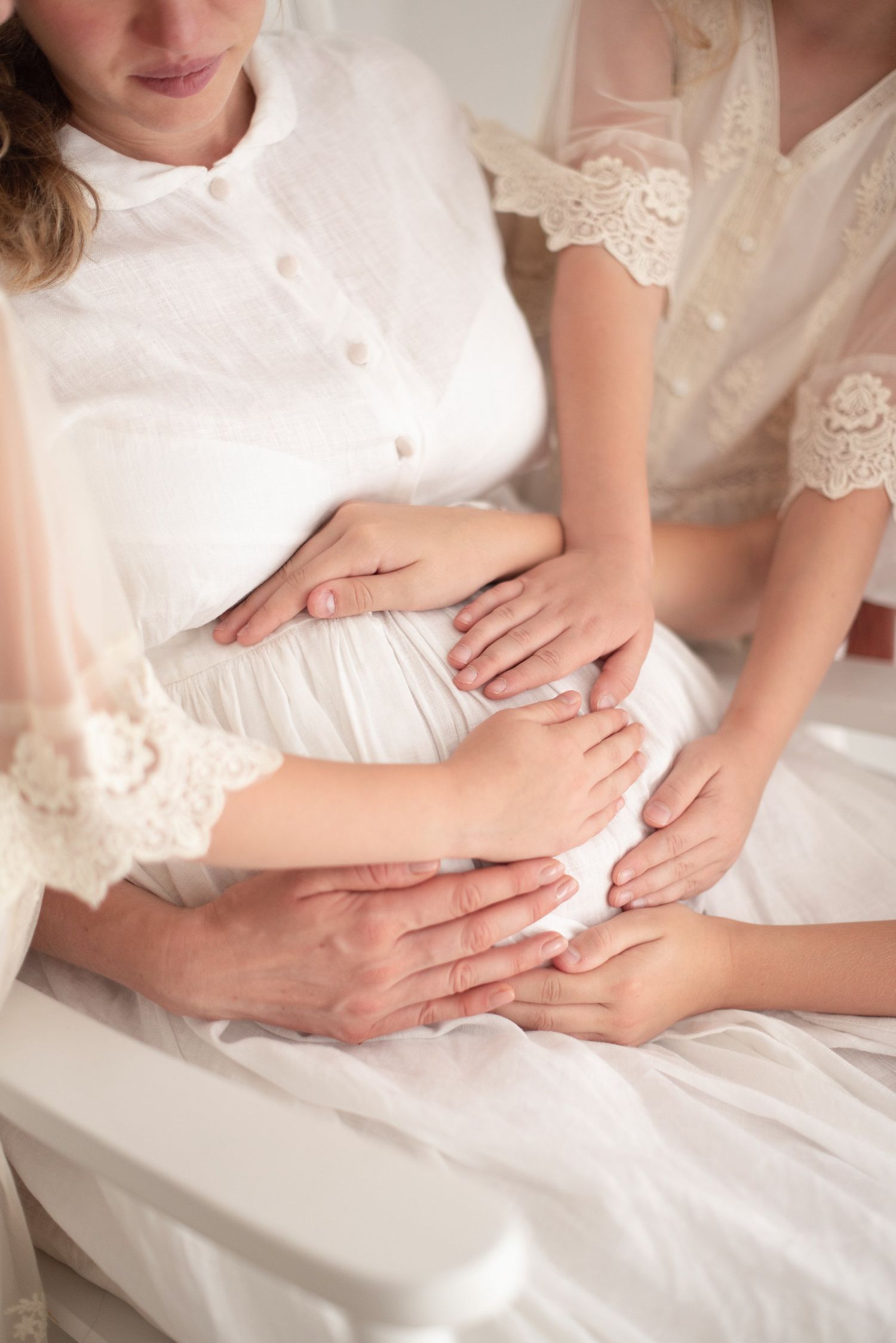 Multiple hands rest on a pregnant belly covered in white lace dress.
