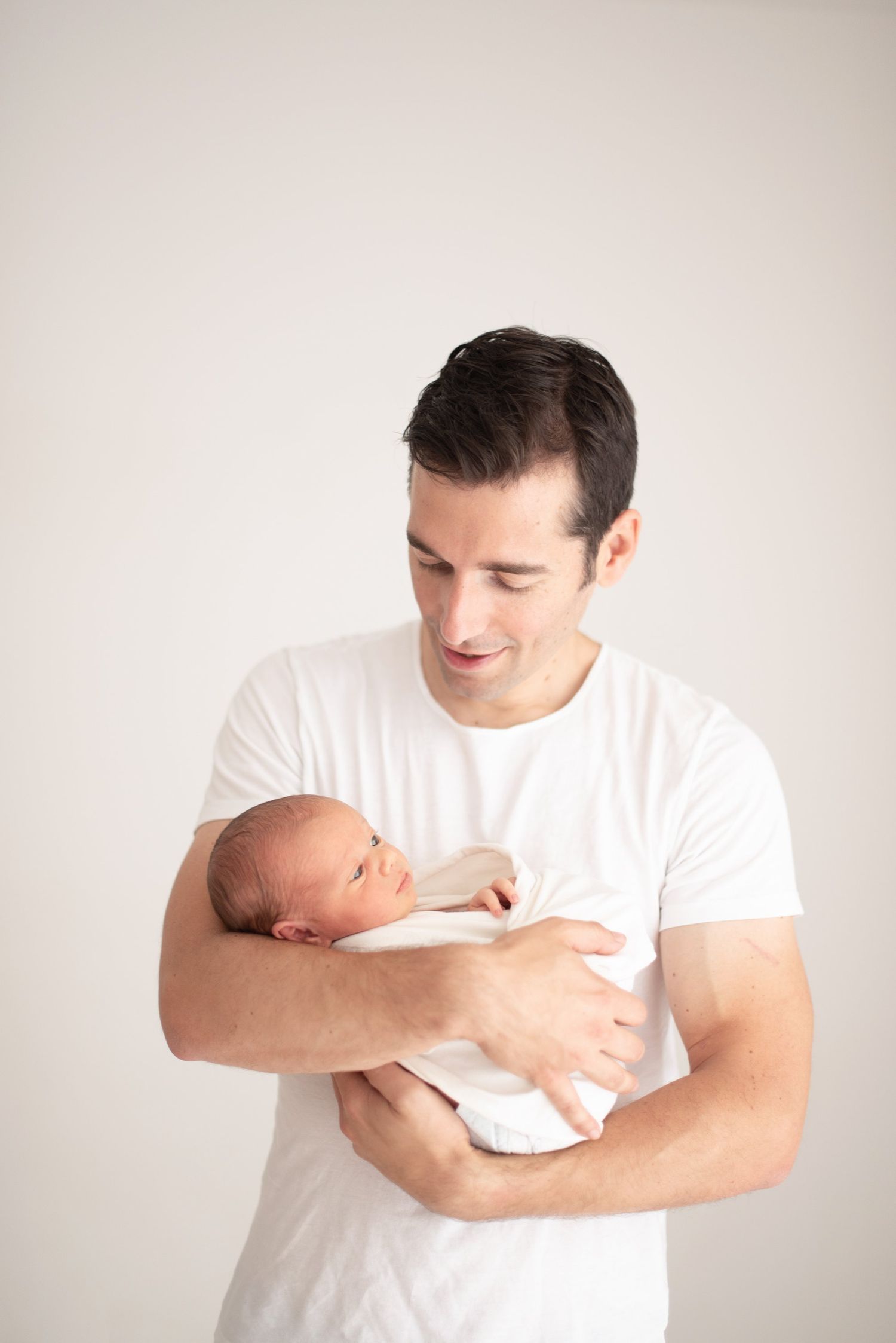 Parent in white shirt holding sleeping newborn baby against light background.