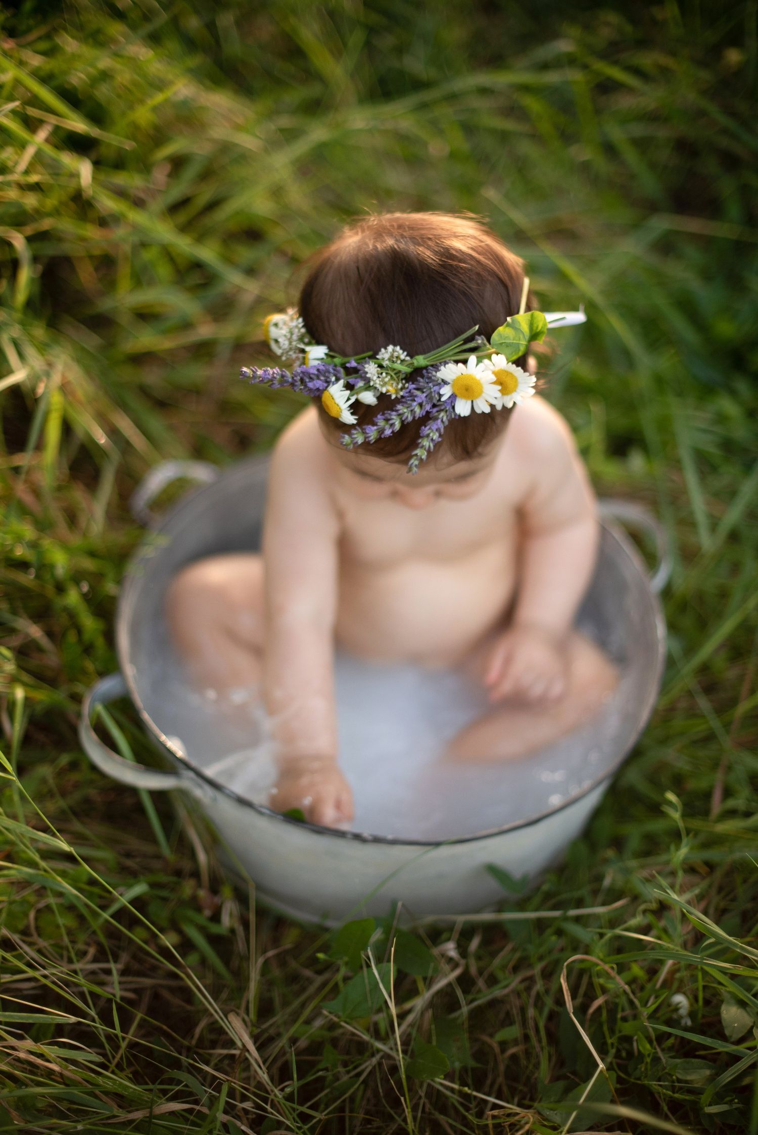 Overhead view of baby in metal tub wearing wildflower crown surrounded by grass.