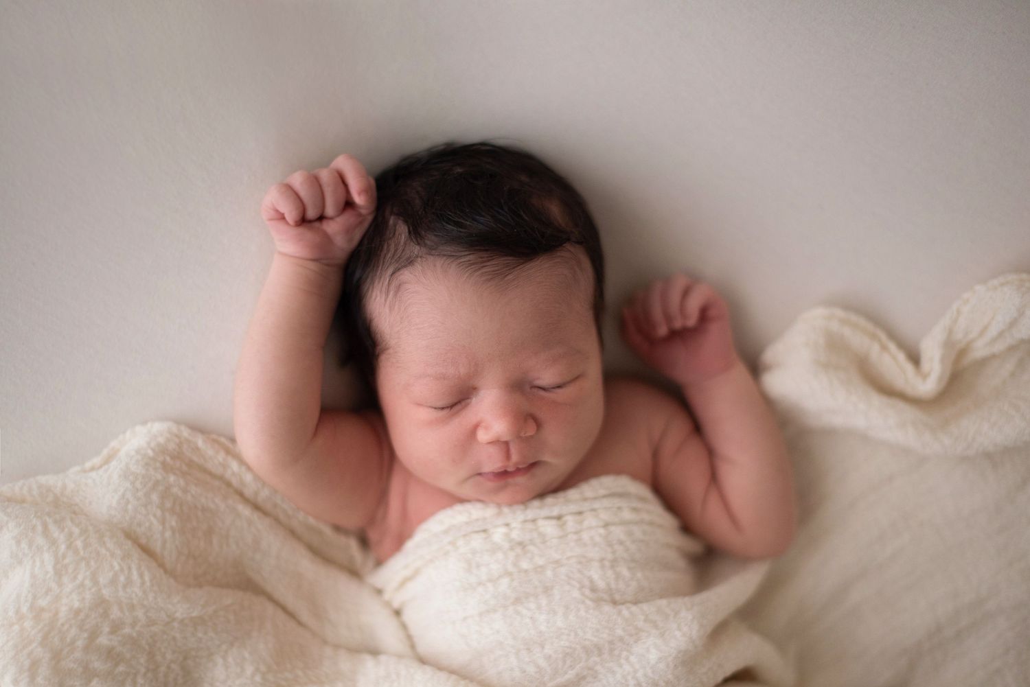 Sleeping newborn wrapped in white blanket with arms raised during studio photo session.
