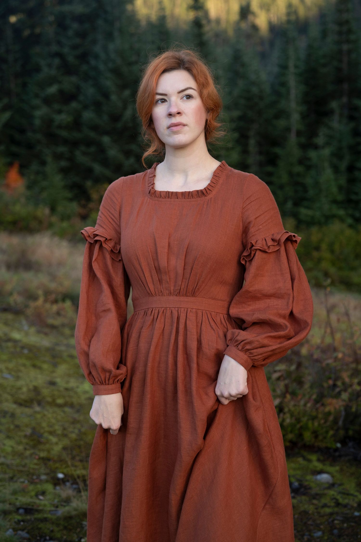 A rustic portrait featuring someone in a ruffled brown prairie dress with balloon sleeves against evergreen trees.