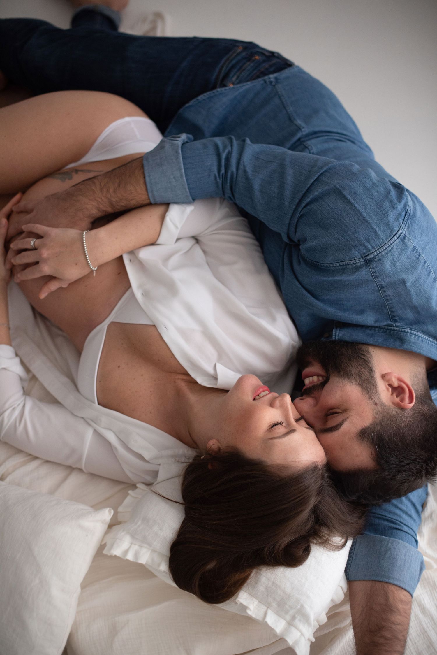 An intimate couple dressed in white and denim share a romantic moment on white bedding.