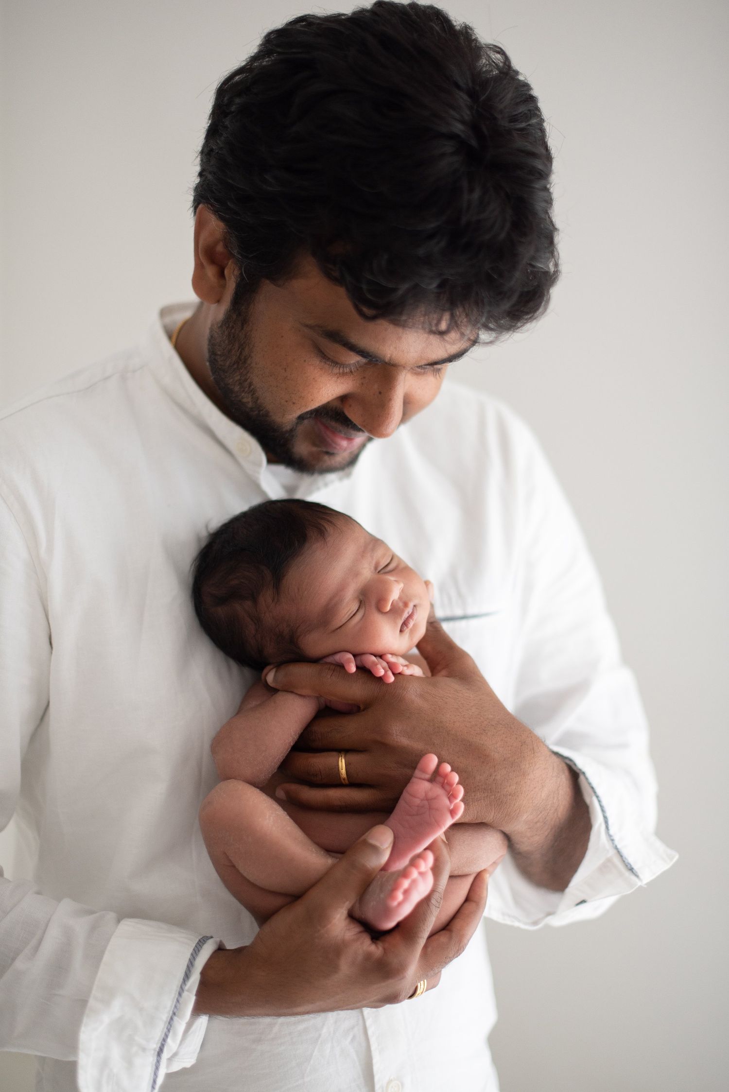 Father in white shirt tenderly holds sleeping newborn baby against light backdrop.