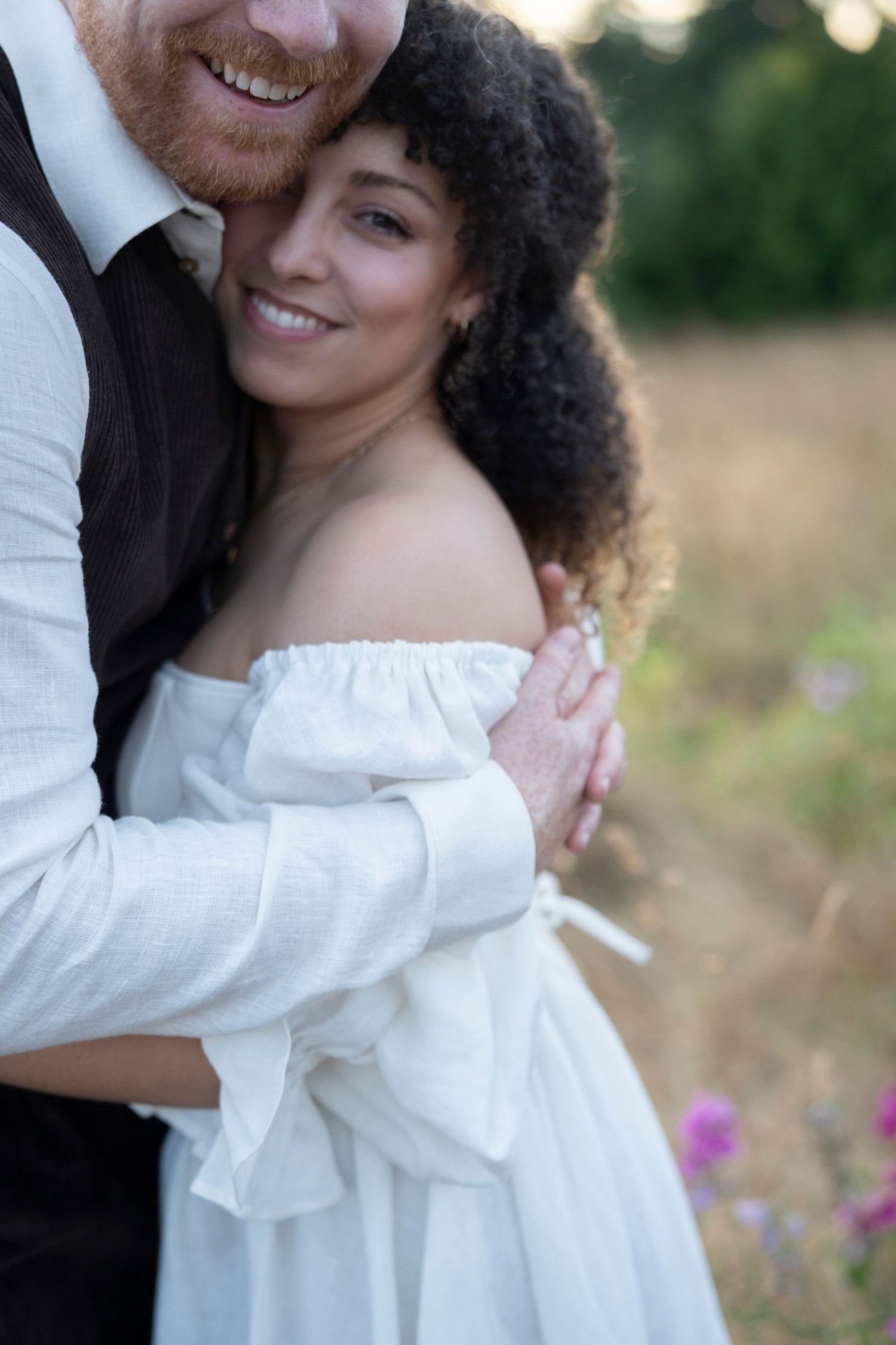A tender embrace between a couple outdoors, with the bride wearing an off-shoulder white dress in a sunlit field.