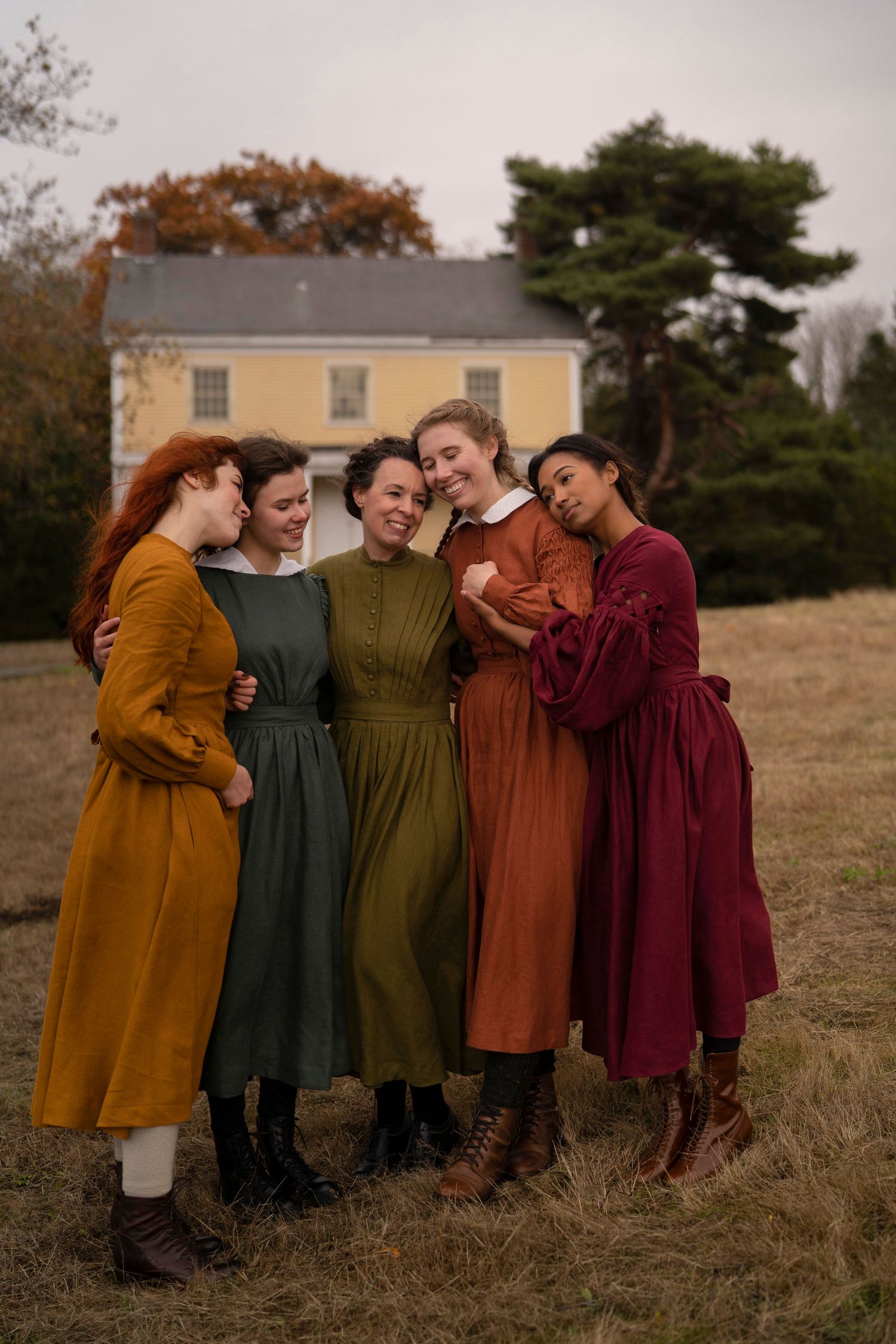 Friends in vintage dresses share a laughing moment together on an autumn field with a farmhouse backdrop.
