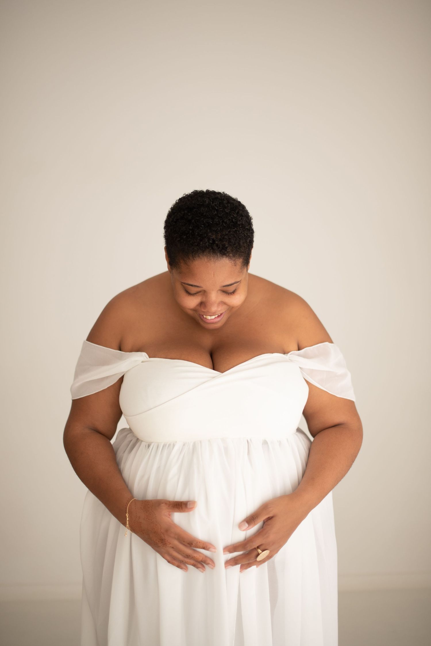 Expectant mother in white off-shoulder dress smiles while cradling baby bump against neutral background.