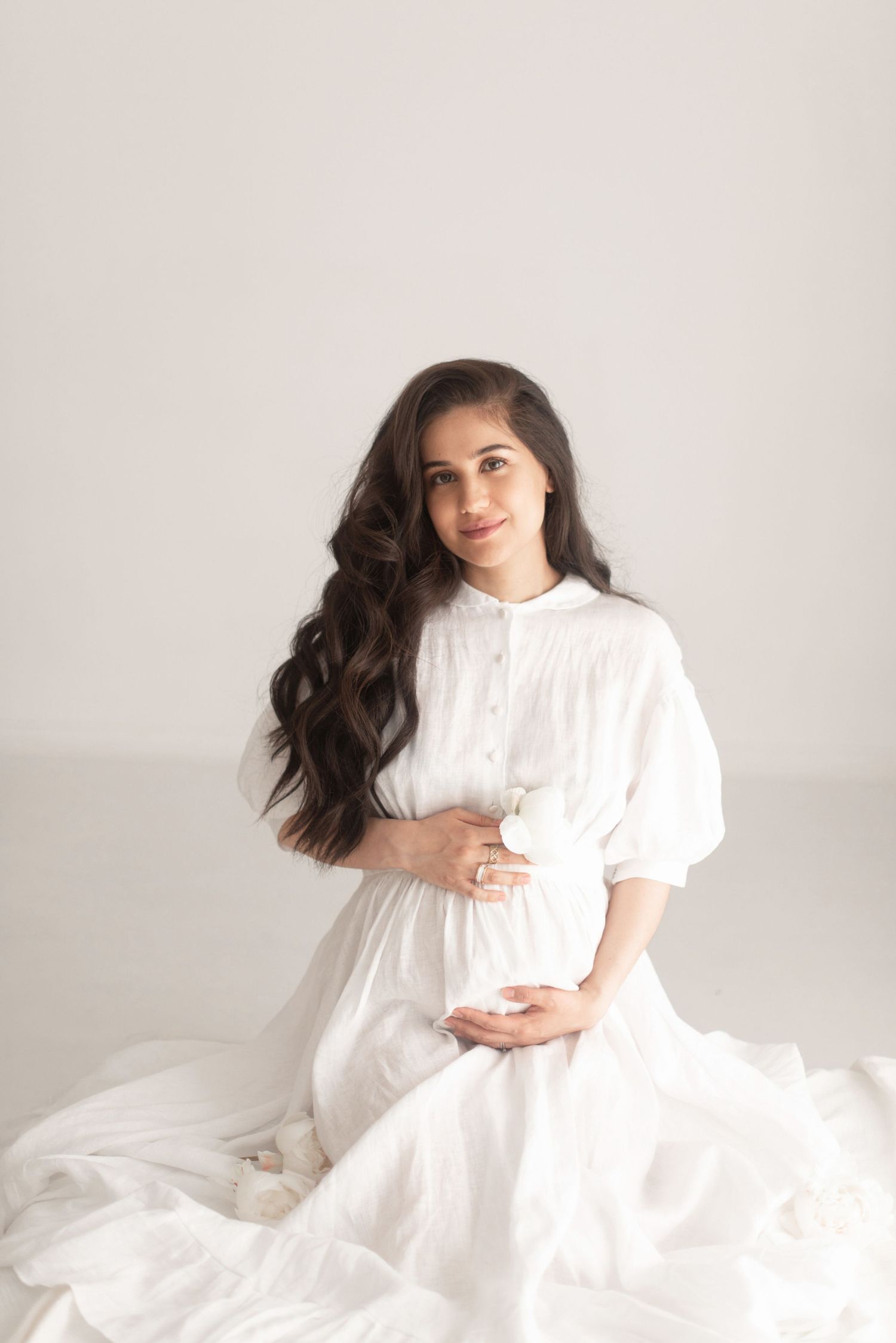 Woman in white dress with long dark hair poses gracefully for maternity portrait.