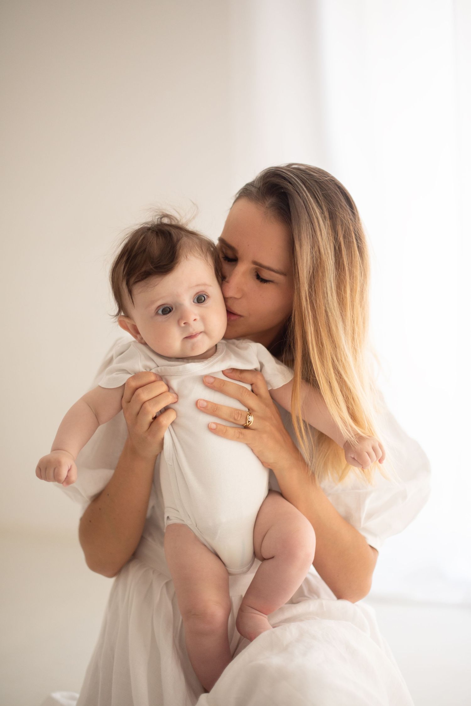 Tender motherly moment between parent and baby in white clothing against light background.