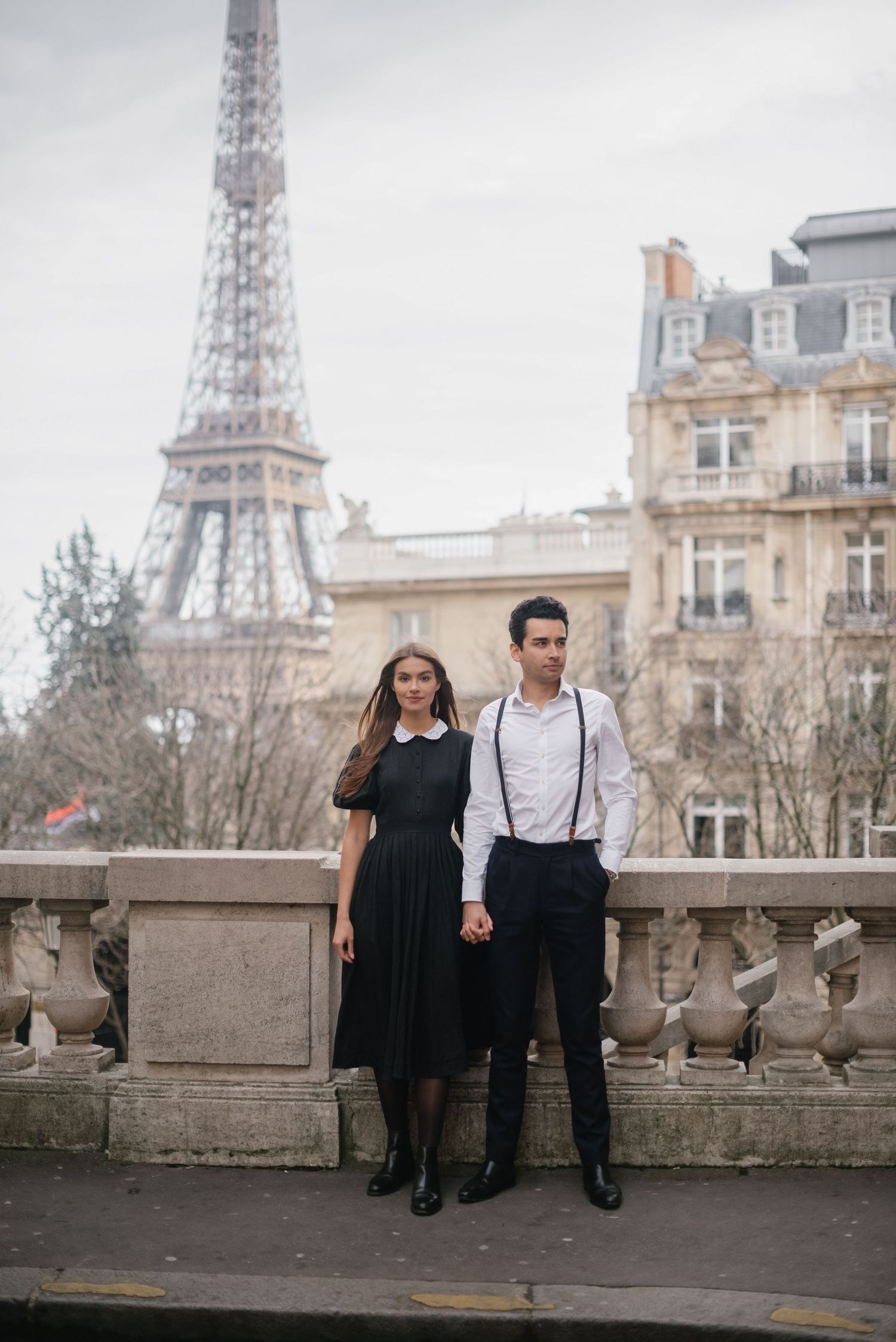 A stylish couple poses on a Parisian balcony with the Eiffel Tower and classic architecture in the background.