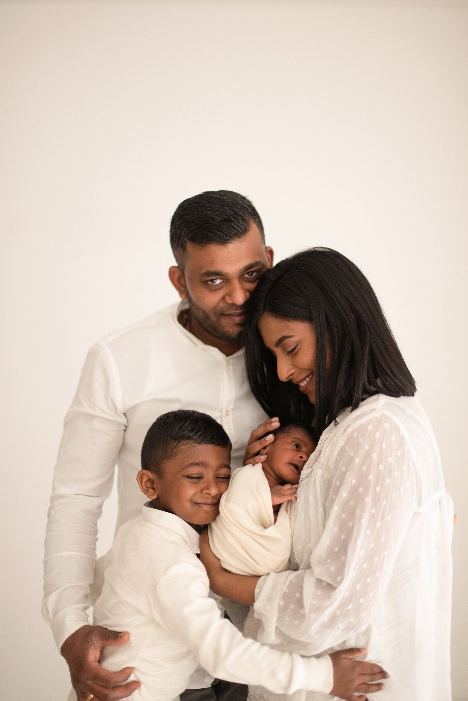 Family of four dressed in white embraces in a tender portrait against neutral background.