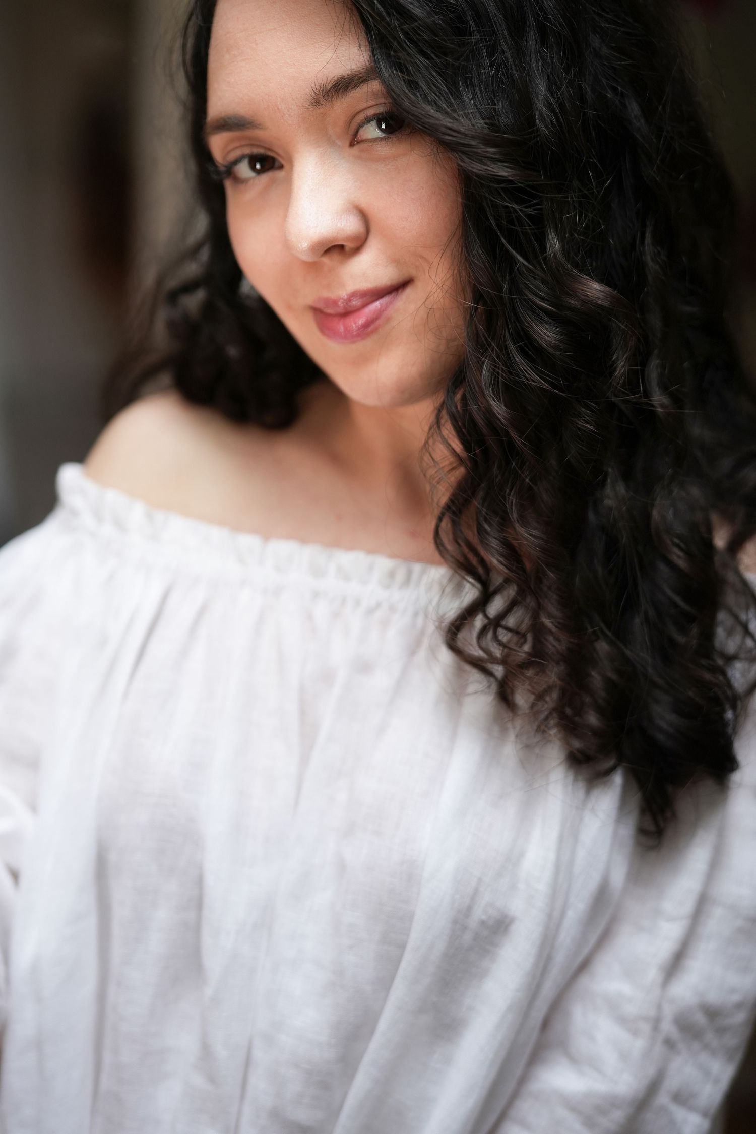 Portrait of someone wearing a white off-shoulder top with long dark curly hair and a gentle smile.