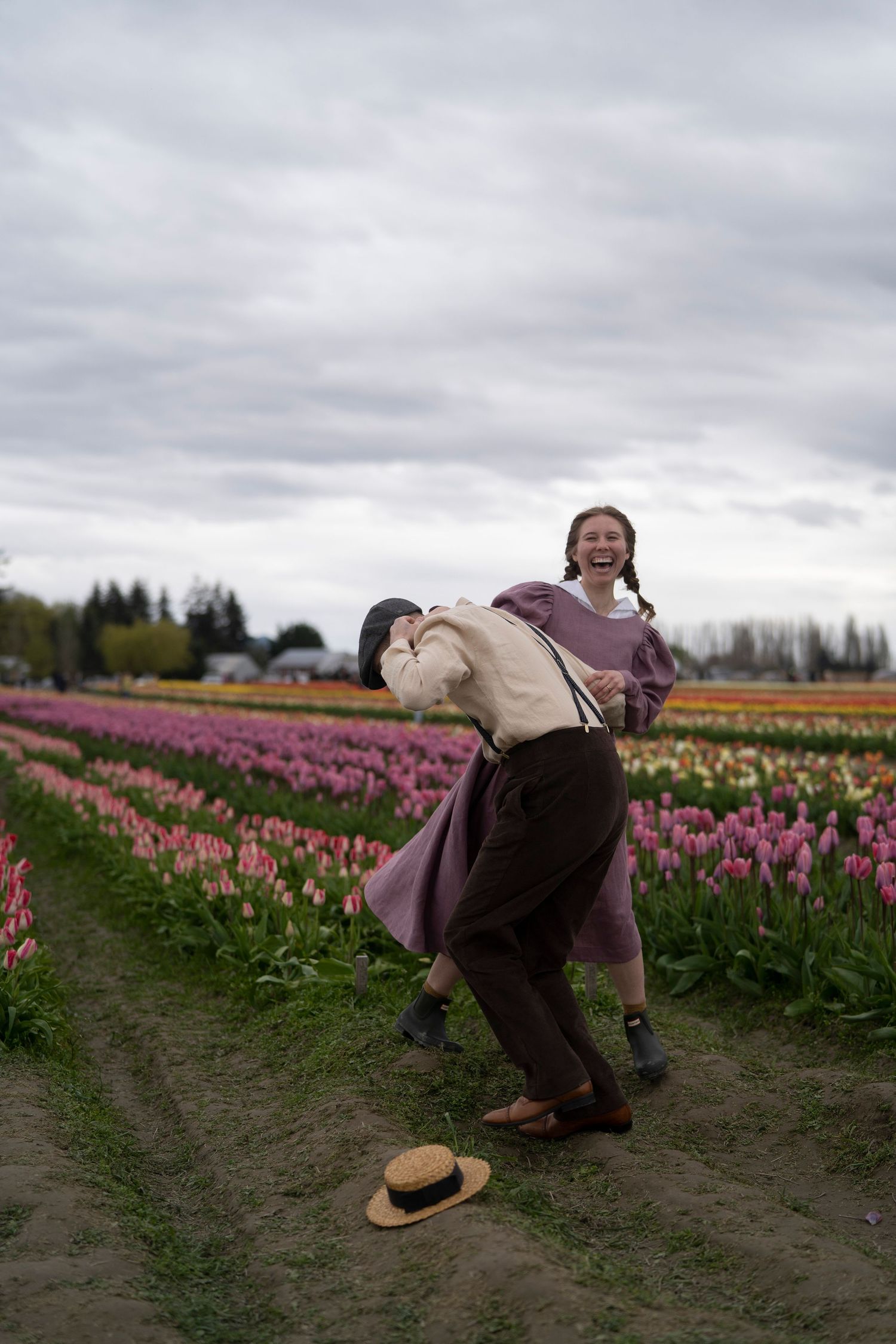 Two people in vintage clothing dancing joyfully between rows of colorful tulips on a cloudy day at a flower farm.