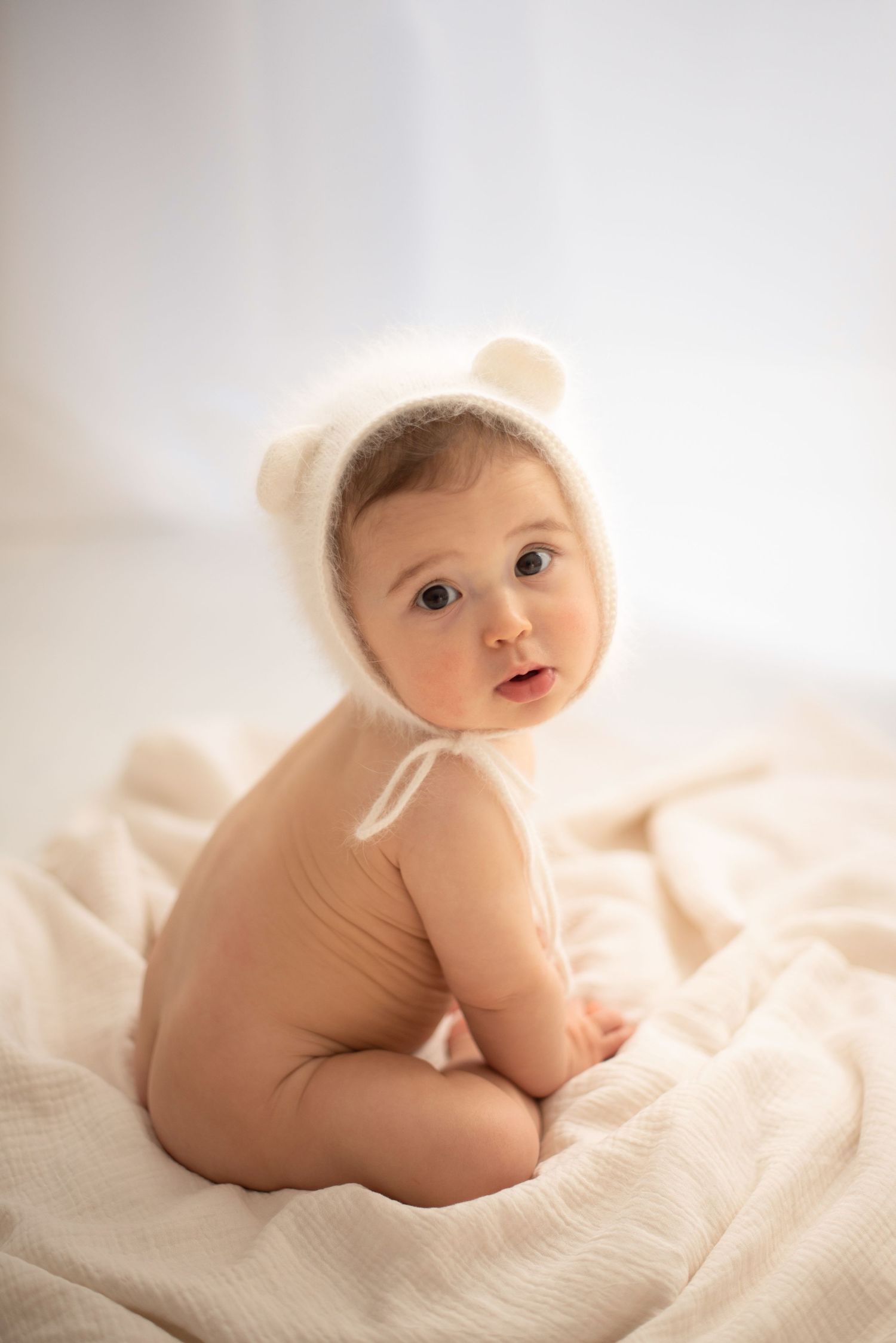 Baby in bear bonnet sitting on cream textile in soft lighting.