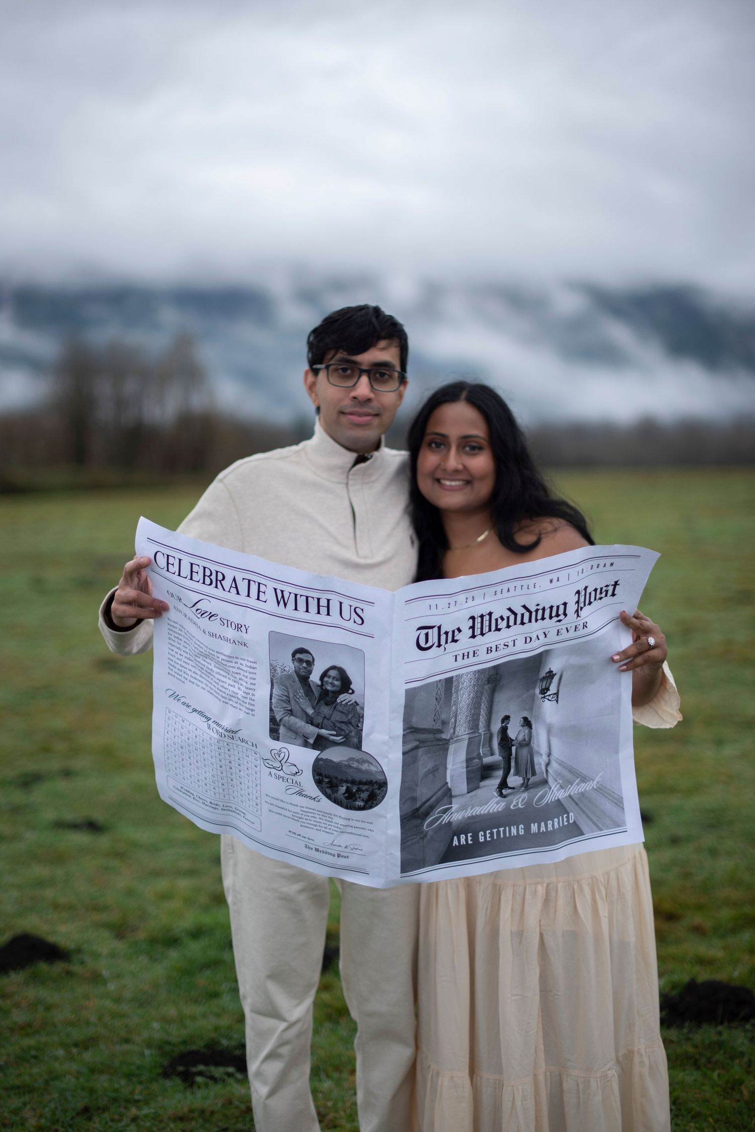 A couple in coordinated outfits holds up a newspaper together in a field on an overcast day.