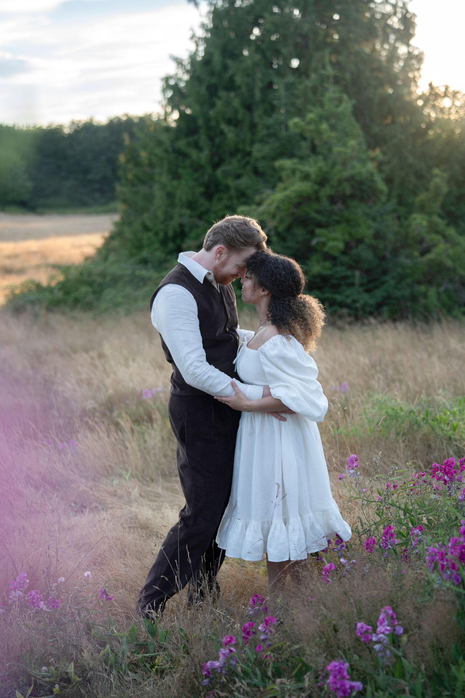 A romantic couple embraces in a field of purple wildflowers at sunset with evergreen trees in the background.