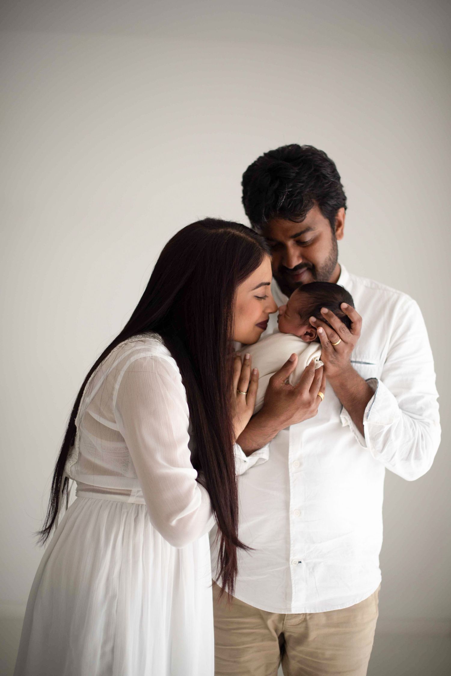 A couple in white outfits sharing a tender moment while gazing down at something together against a neutral backdrop.