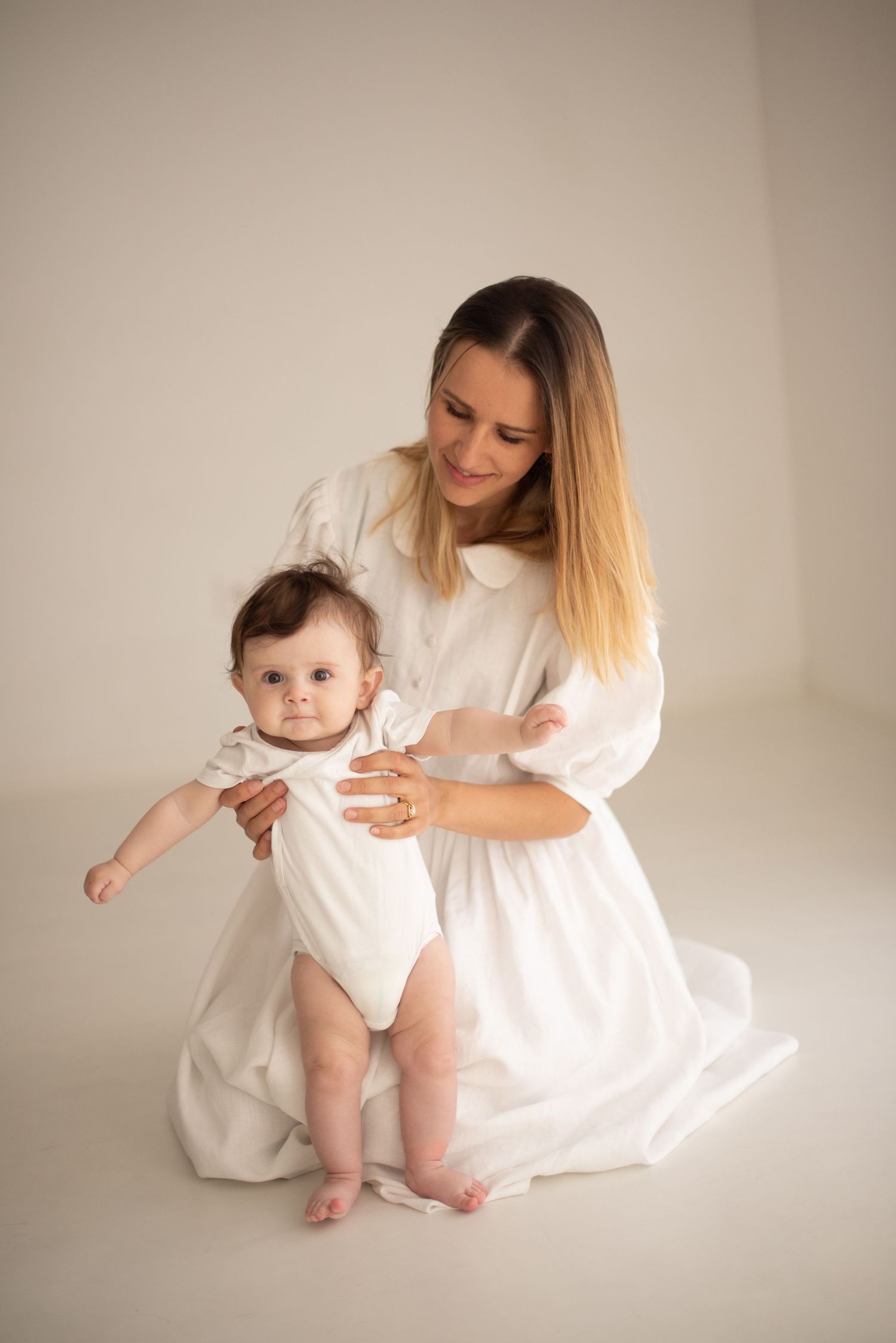 Parent and baby wearing matching white outfits sit together on light studio backdrop during tender portrait session.