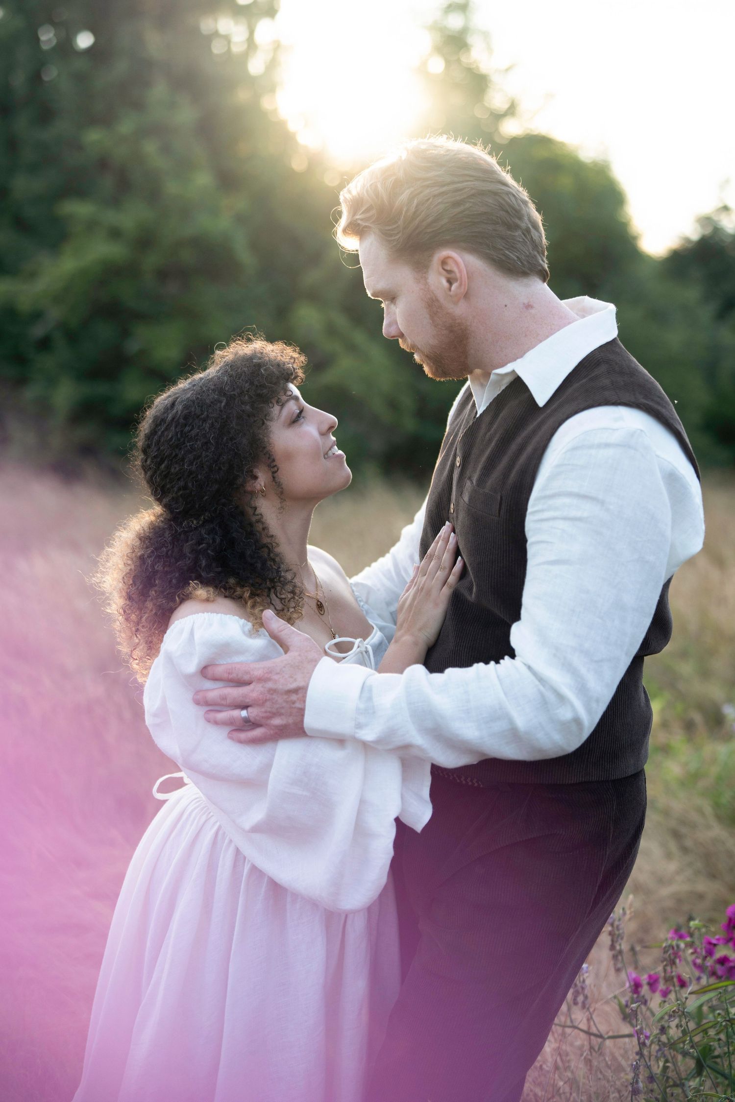 A romantic couple embraces during a sunset photoshoot with pink flowers in the foreground and soft lighting.