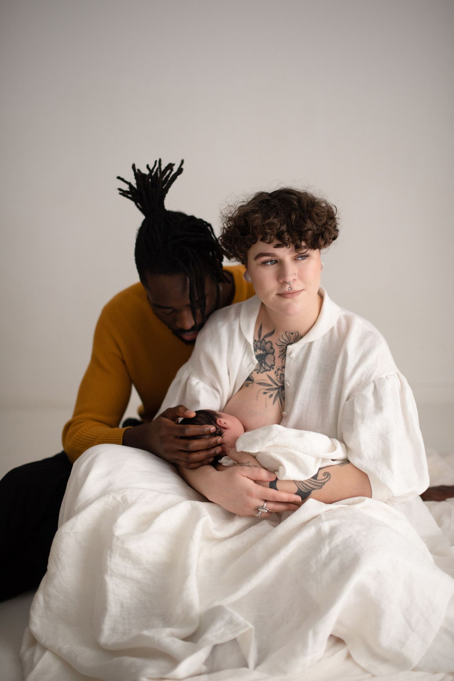 A couple dressed in white and mustard holds a newborn baby in a minimalist studio setting.