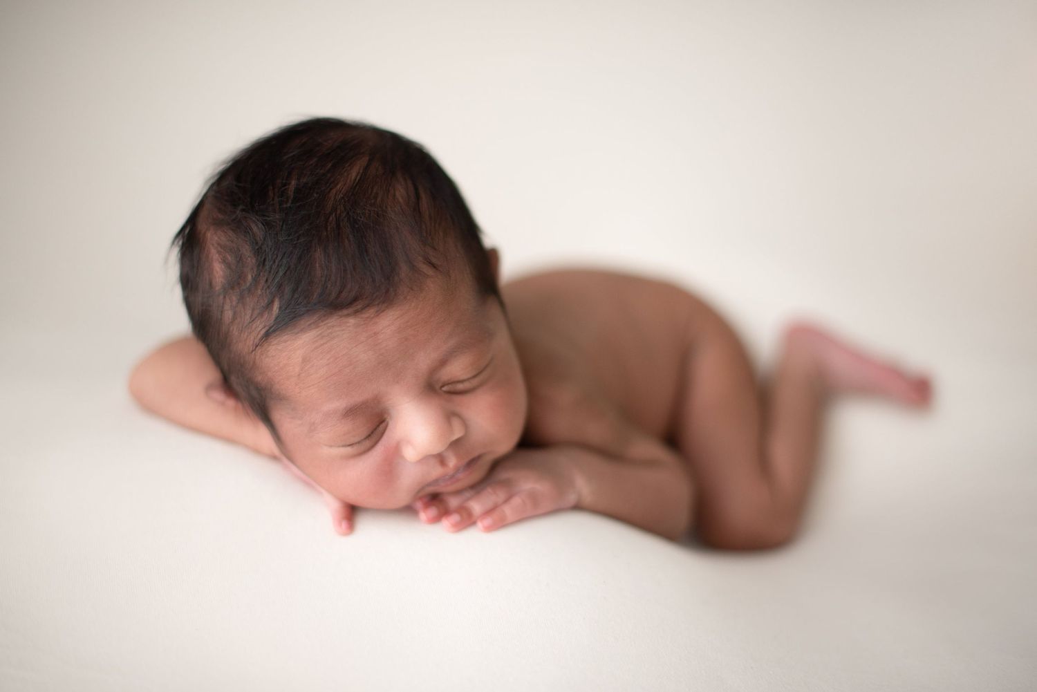 A peaceful sleeping newborn rests their head on folded arms against a soft white background.