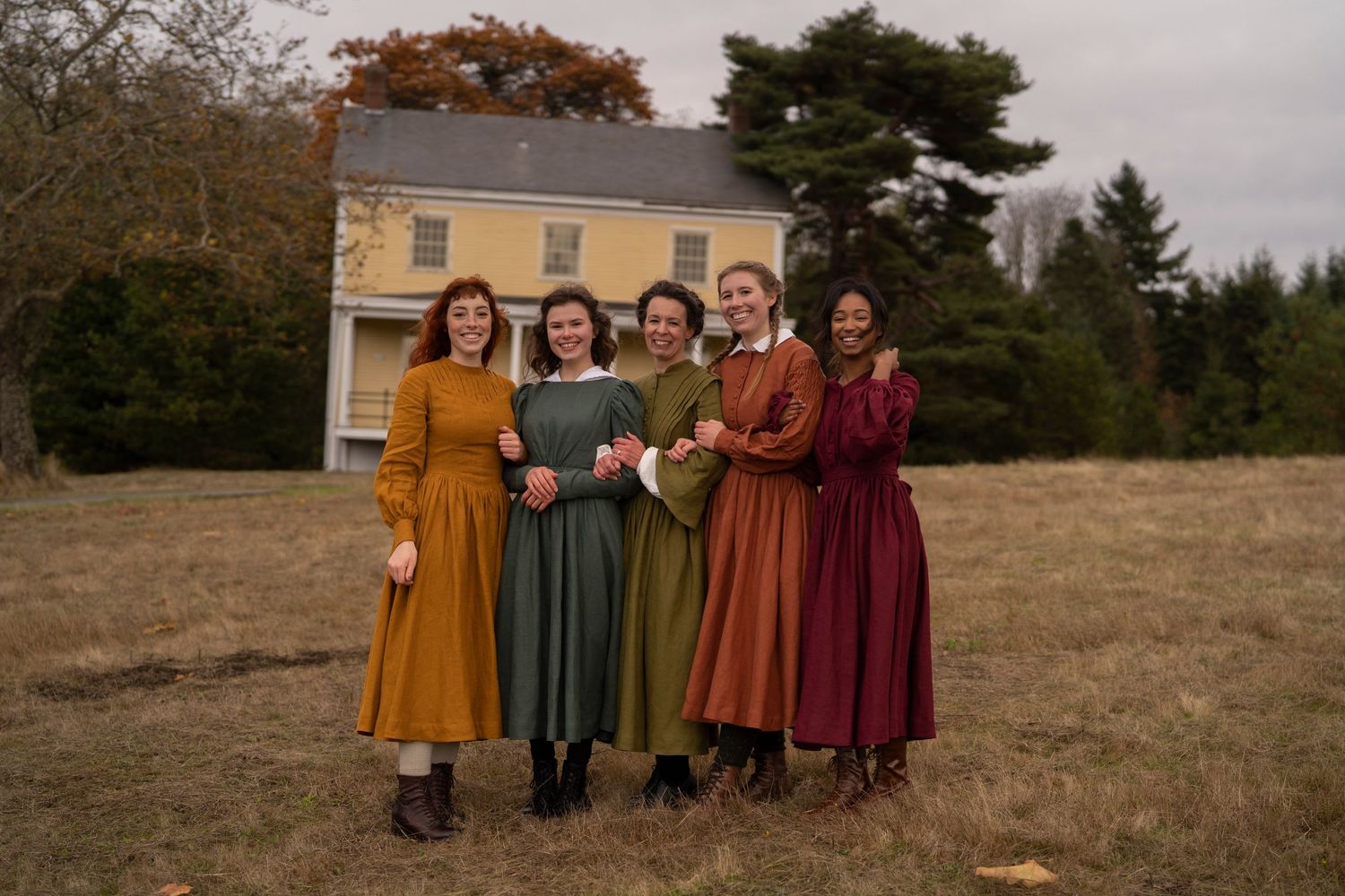 A group in historical dresses in autumnal colors poses together in front of a yellow farmhouse.