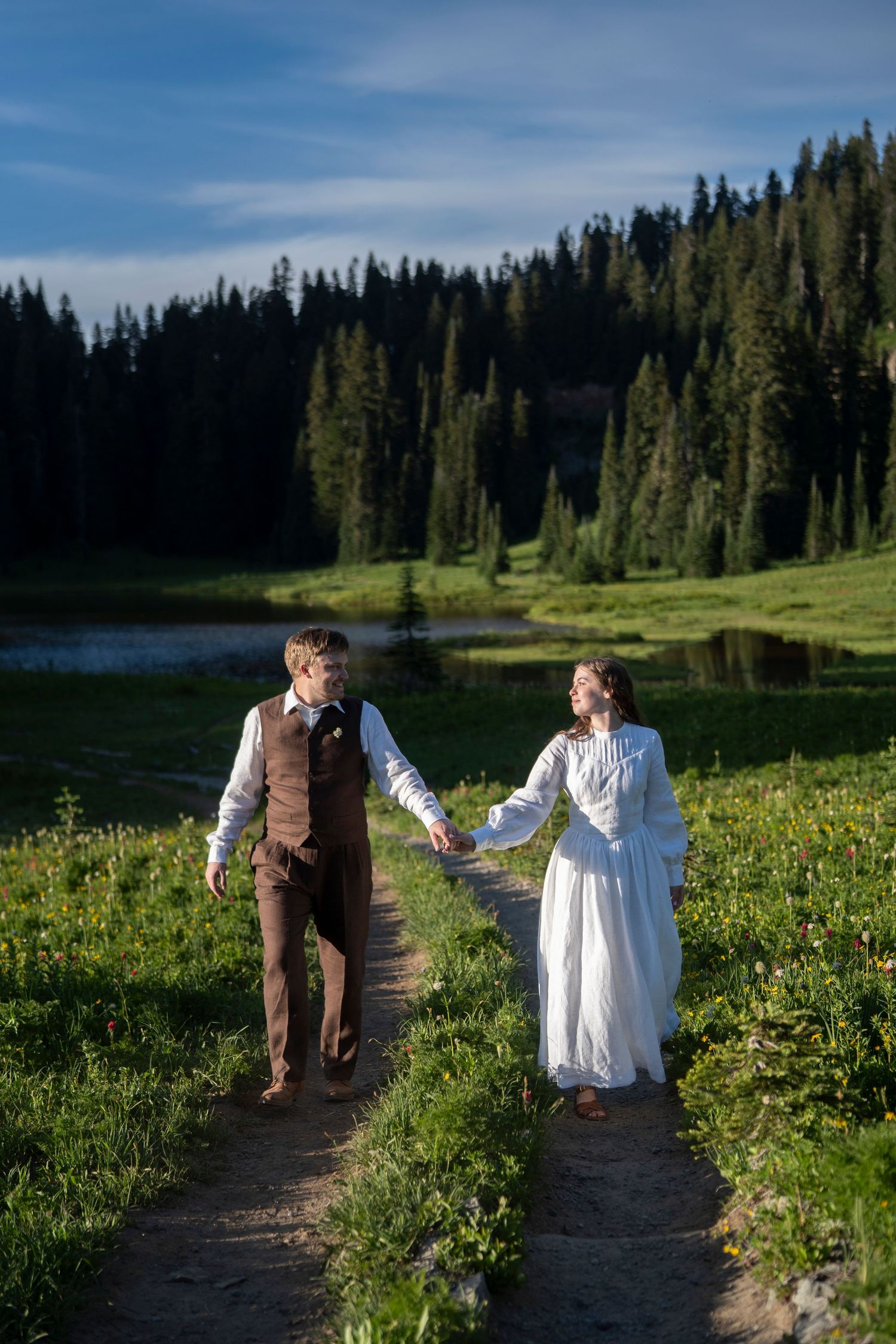 A couple walks together on a mountain trail surrounded by wildflowers and evergreen trees in a scenic landscape.
