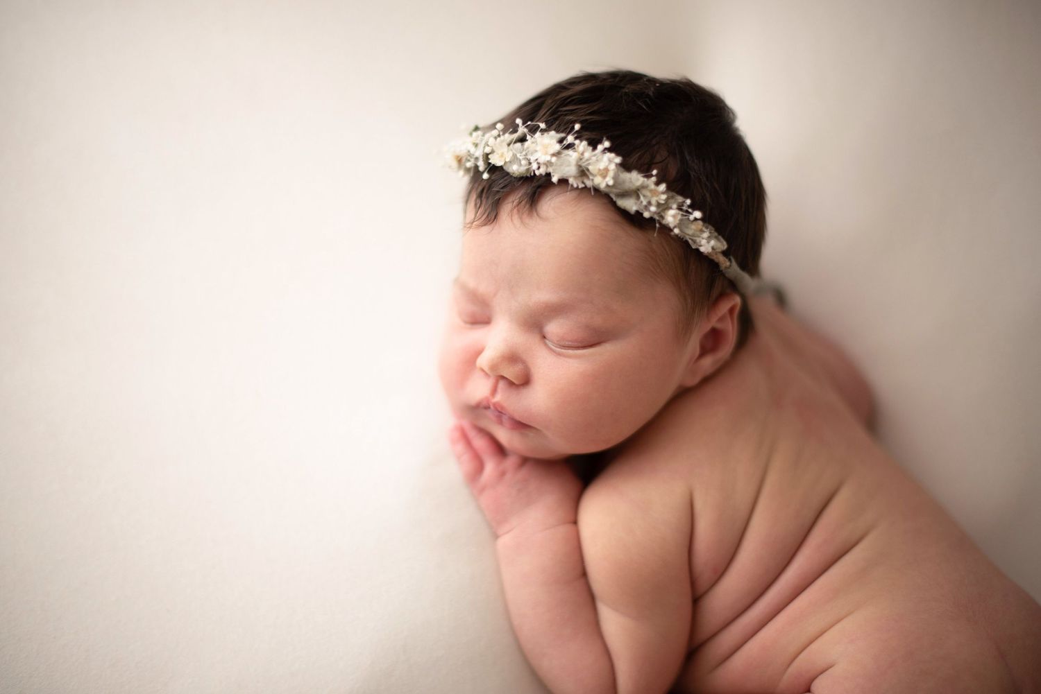 Sleeping newborn wearing delicate floral crown laying on cream backdrop during professional photo session.