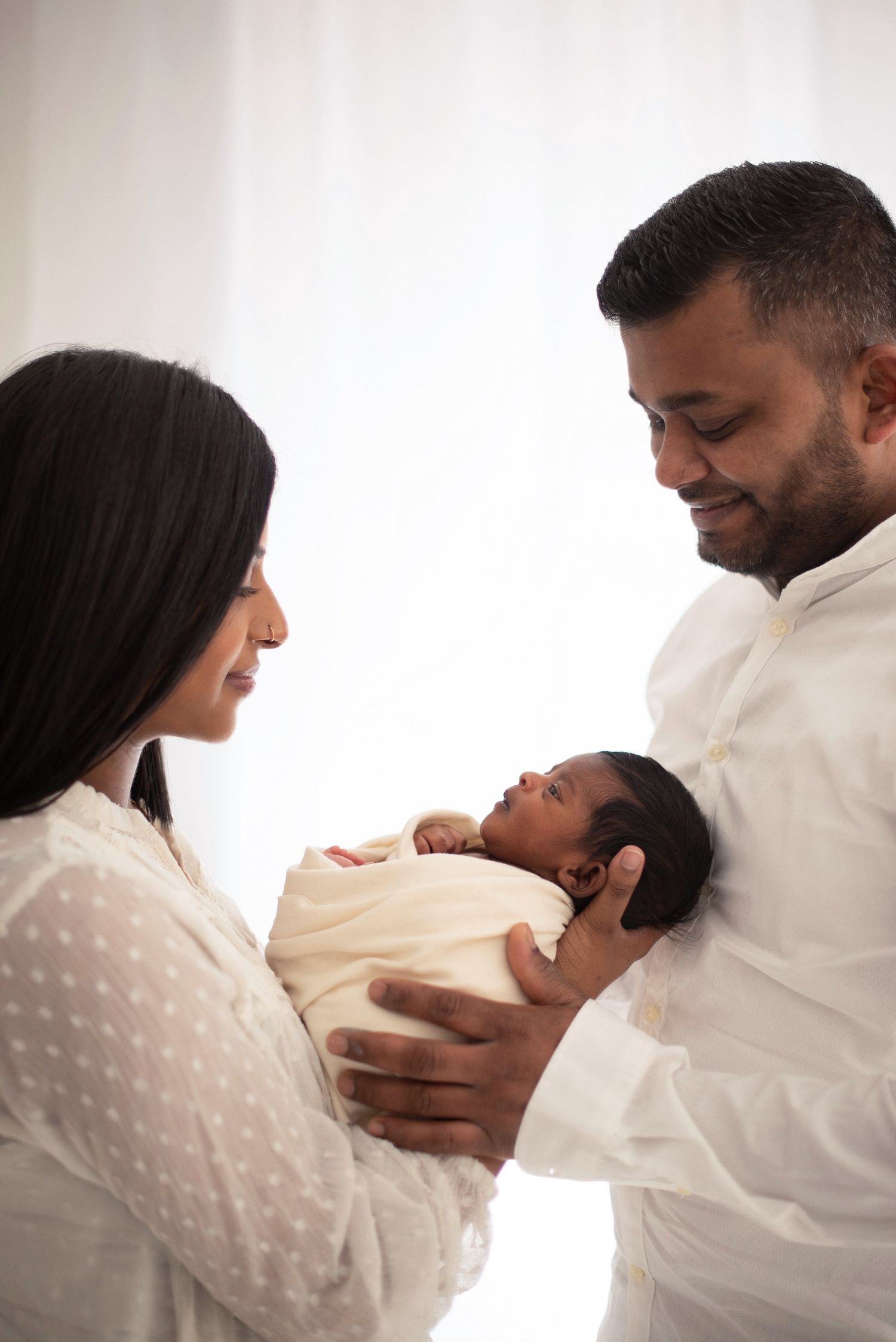 Family members share a loving moment while holding their newborn baby wrapped in soft cream fabric.