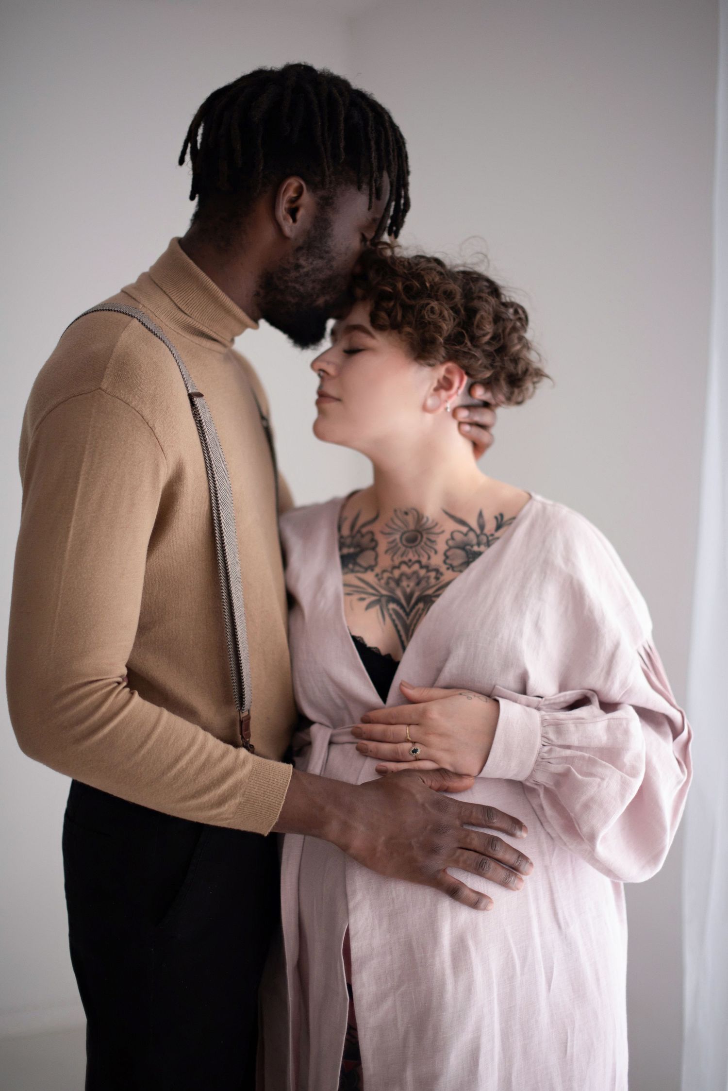 Intimate portrait of expecting couple in neutral tones, sharing gentle moment against light background.