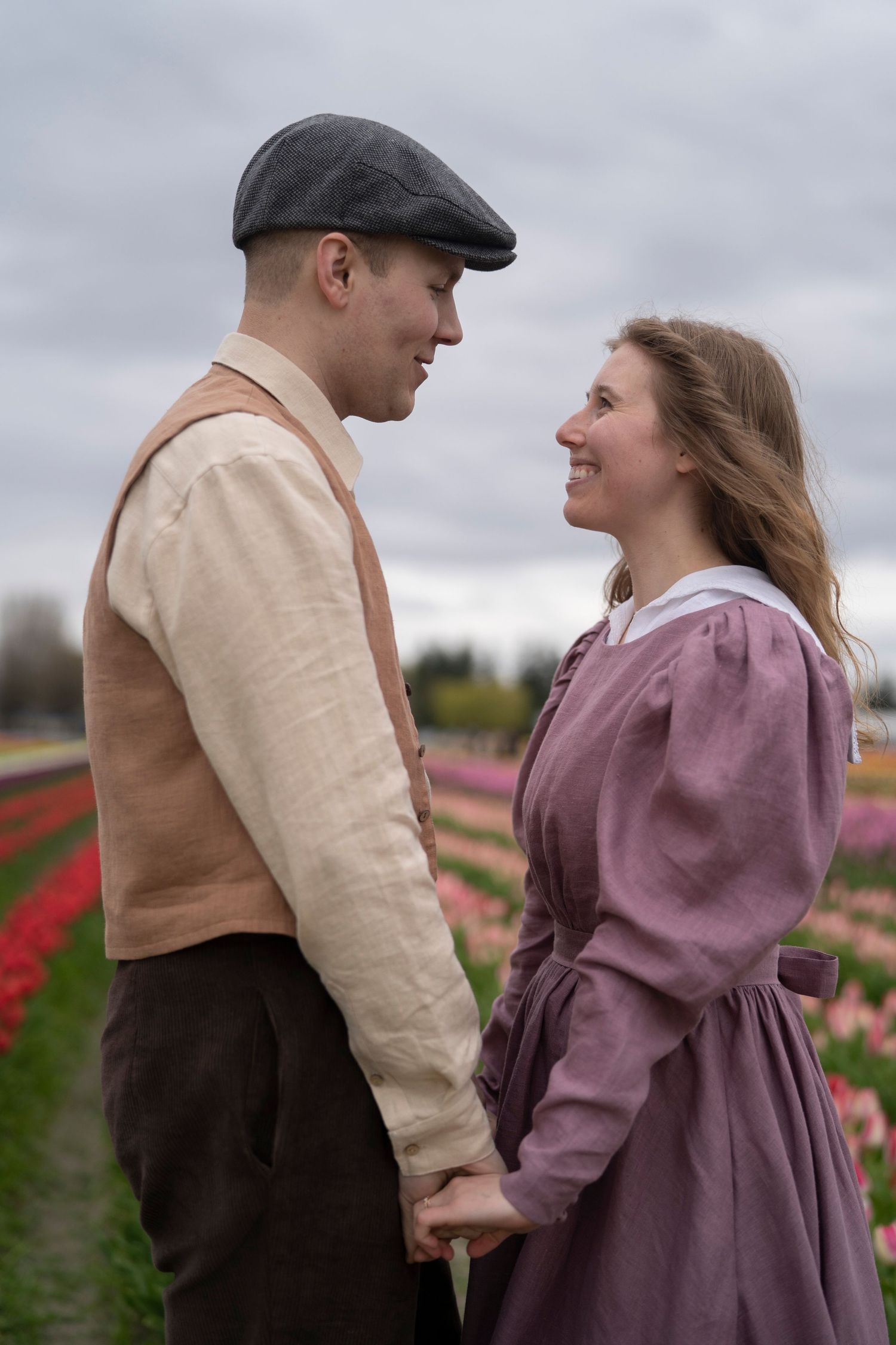A romantic vintage-style couple holds hands while standing in a blooming tulip field under an overcast sky.