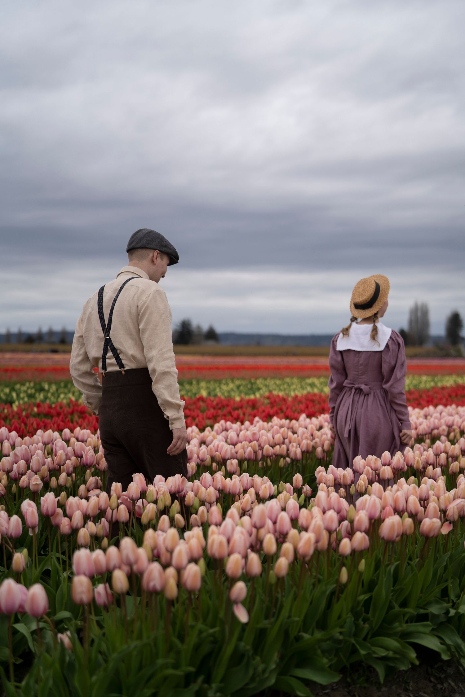 Two people in vintage clothing stand among pink and red tulip fields under a moody cloudy sky.