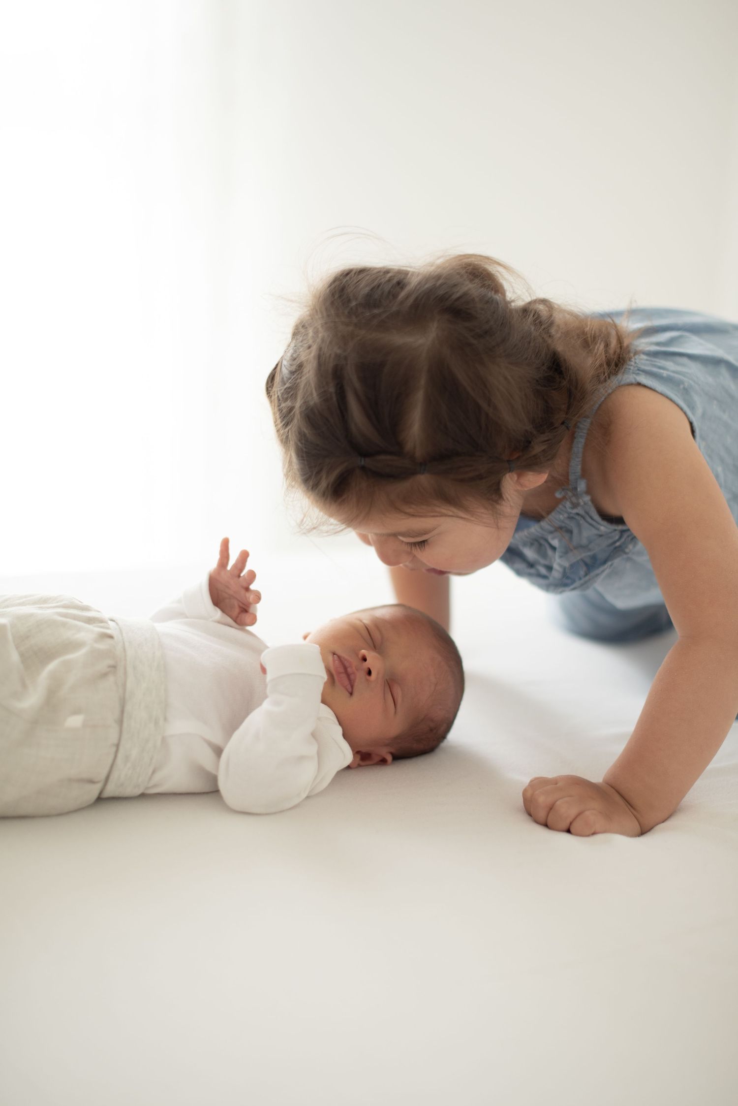 Young child leaning over to interact with newborn baby lying on white surface.