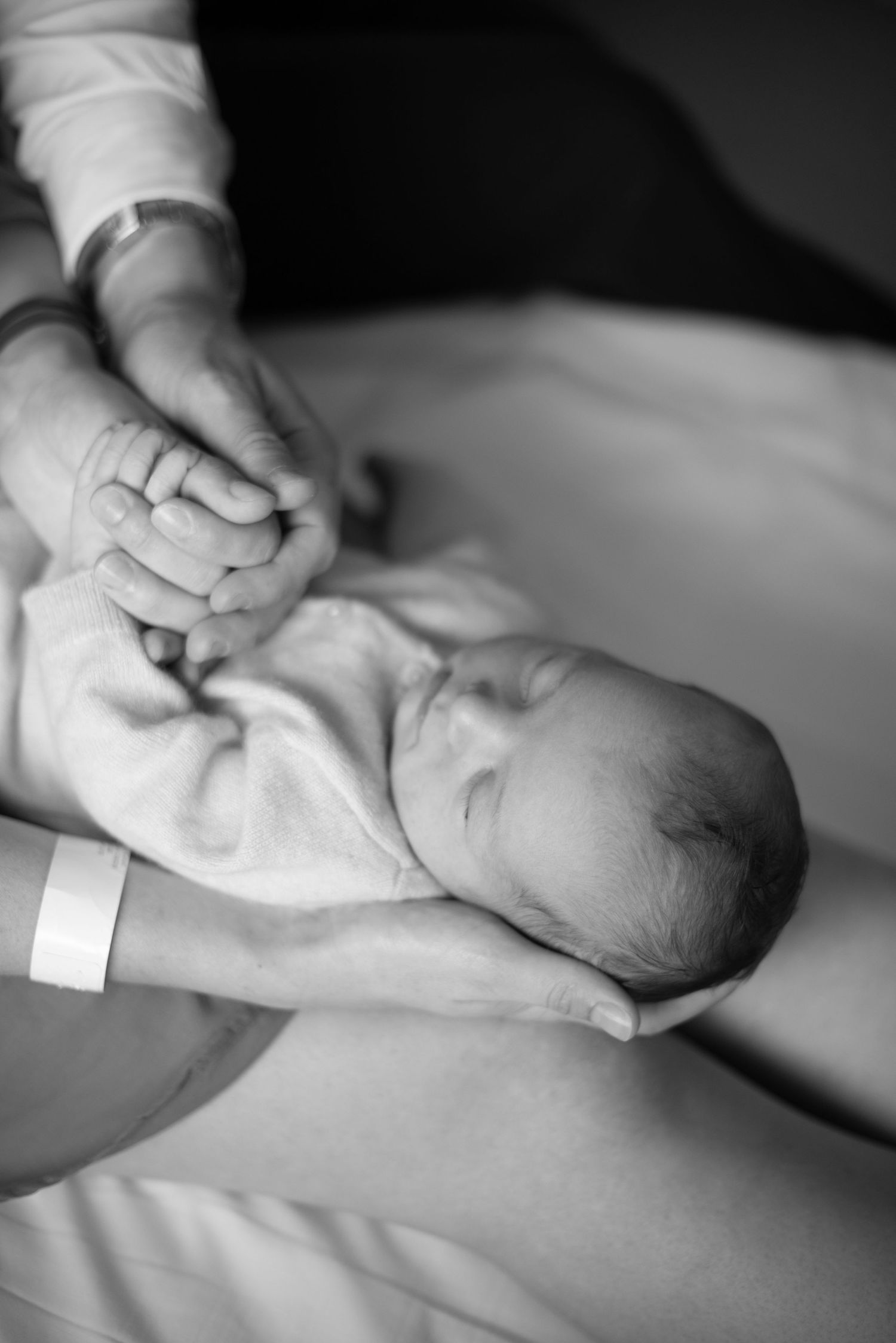 Black and white image of tiny newborn baby's hand being held as they rest in hospital blanket with ID bracelet.