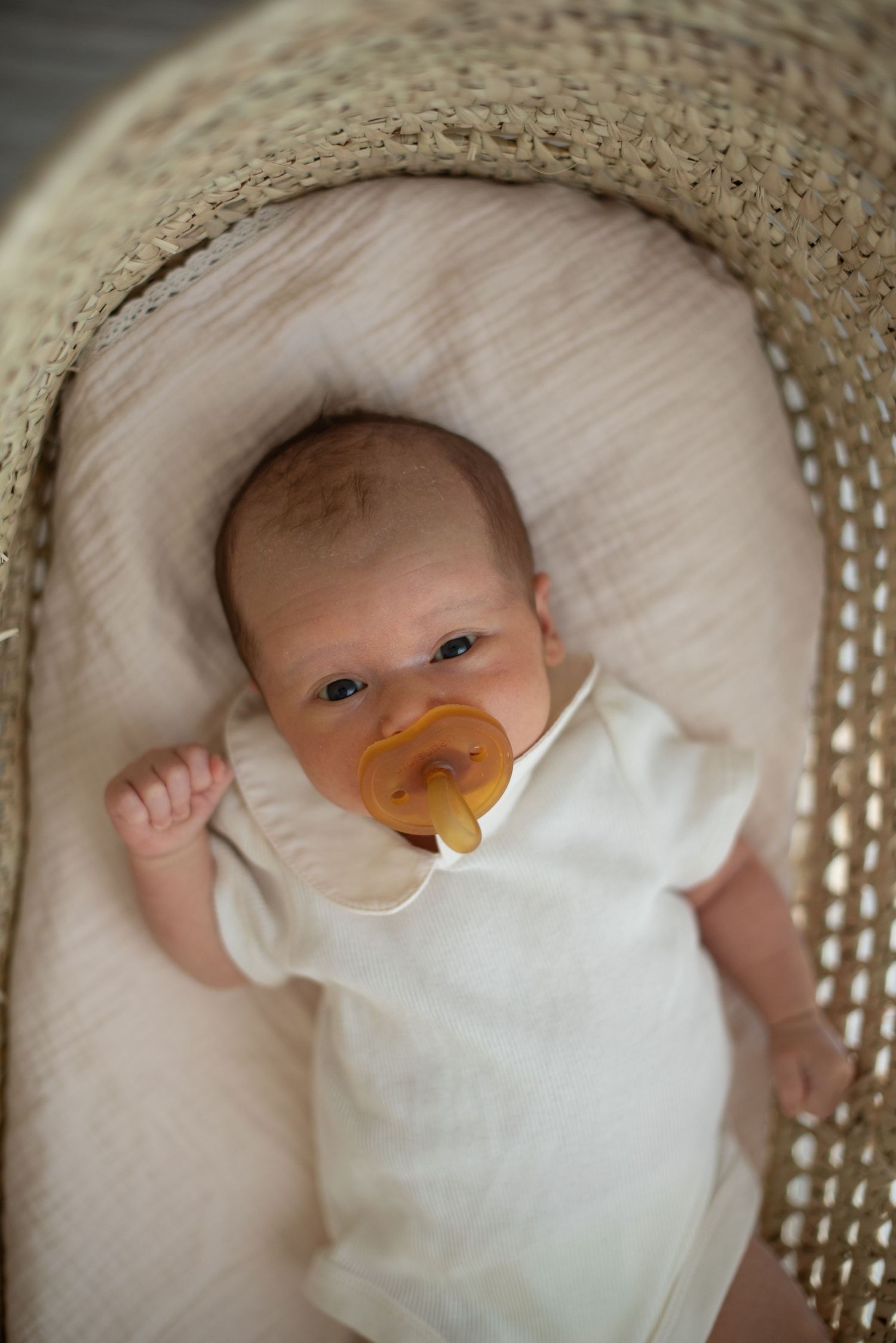 Baby with pacifier resting in woven bassinet wearing white onesie.