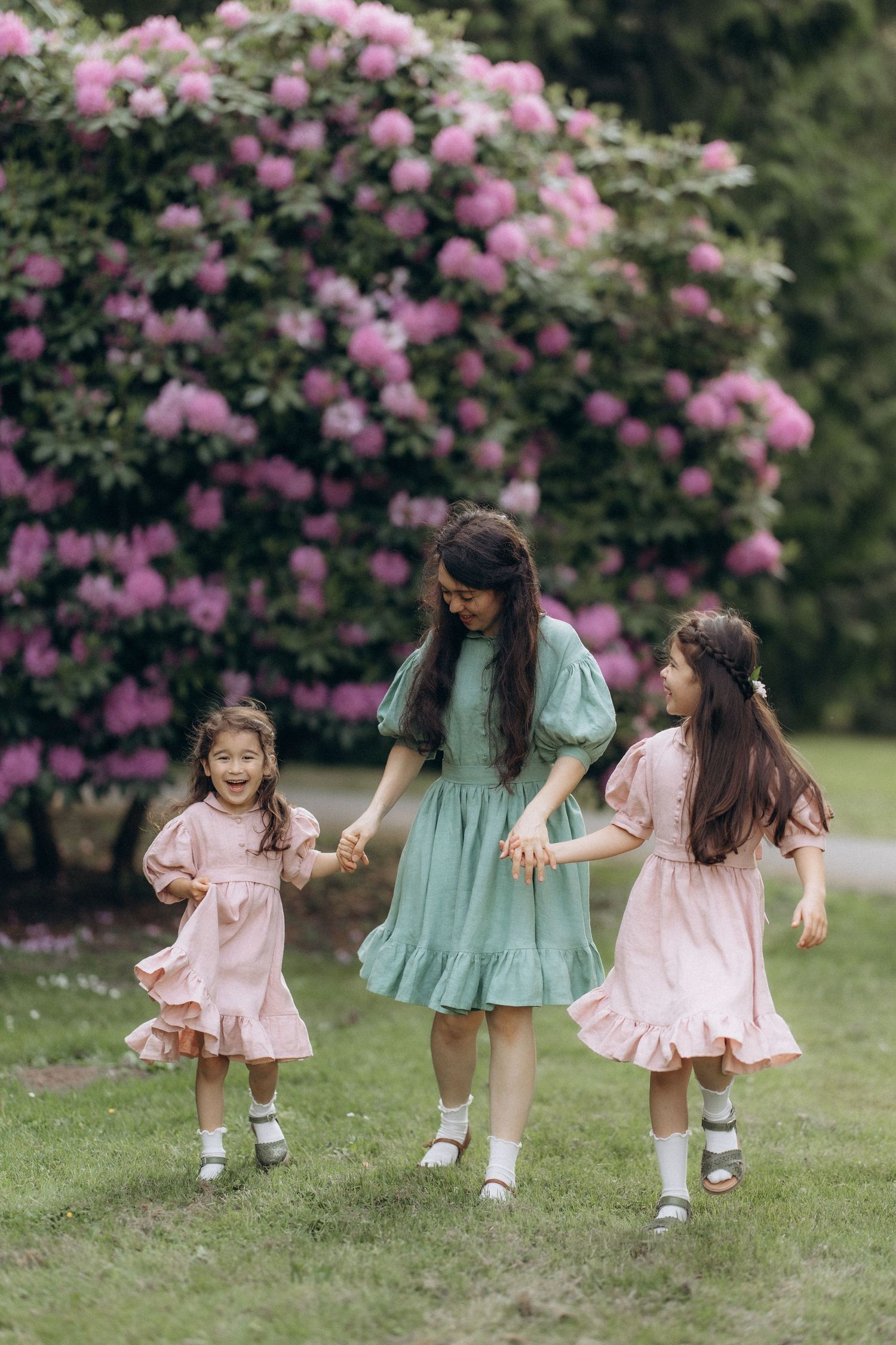 Three people in vintage-style dresses hold hands and skip together on grass near blooming pink rhododendron bushes.