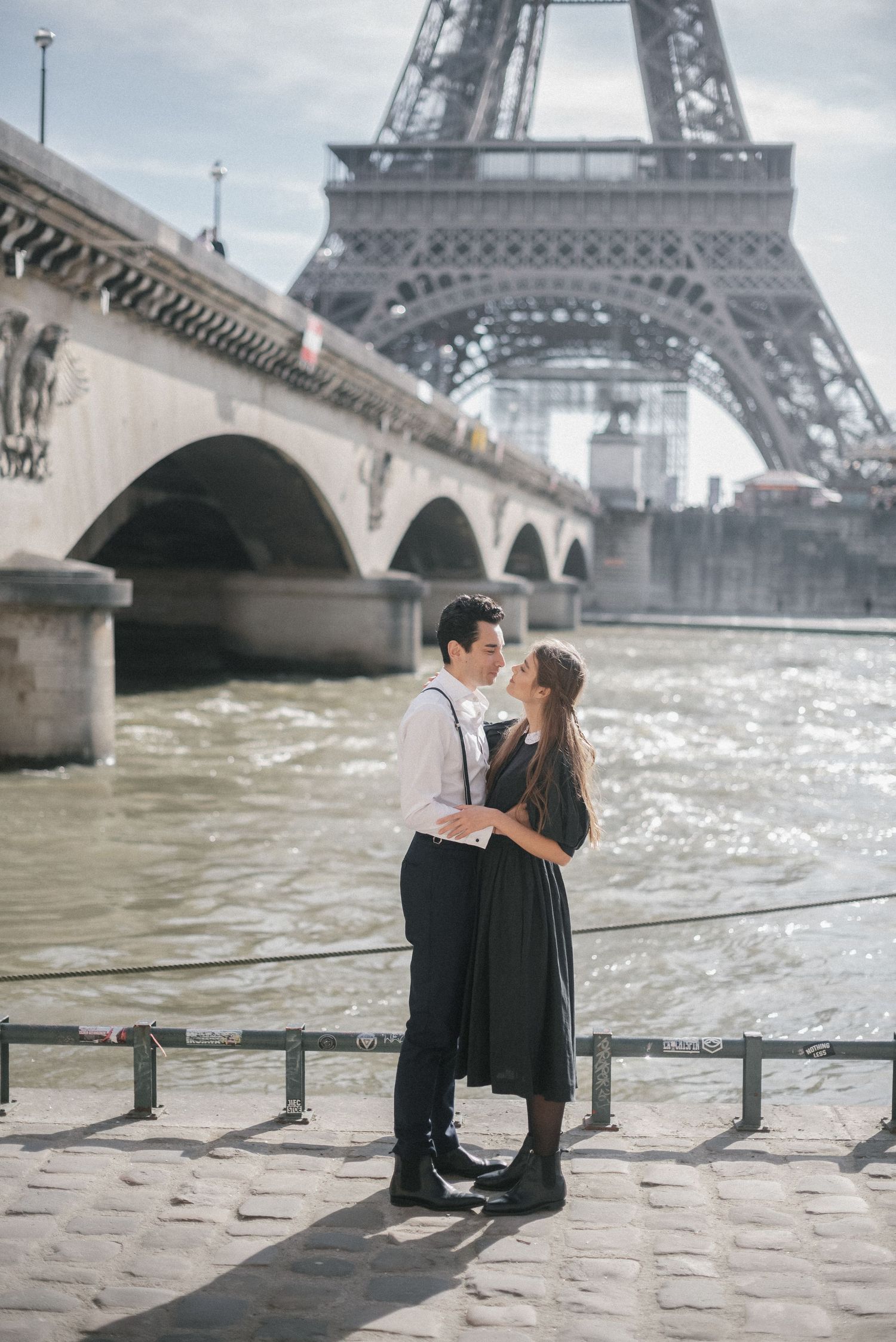 Romantic couple embraces by Seine River with Eiffel Tower in background.