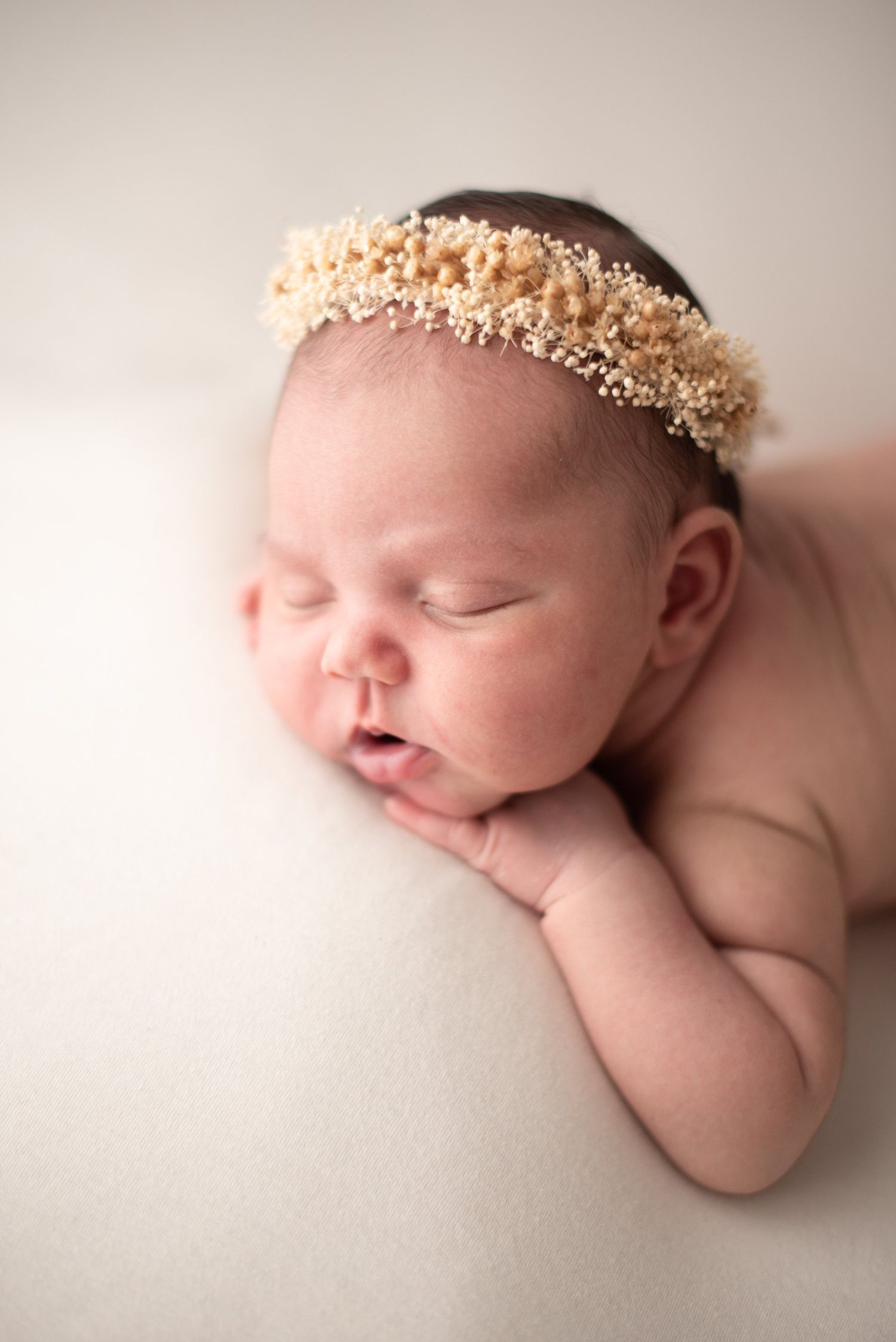 Sleeping newborn wearing delicate floral crown against cream background.