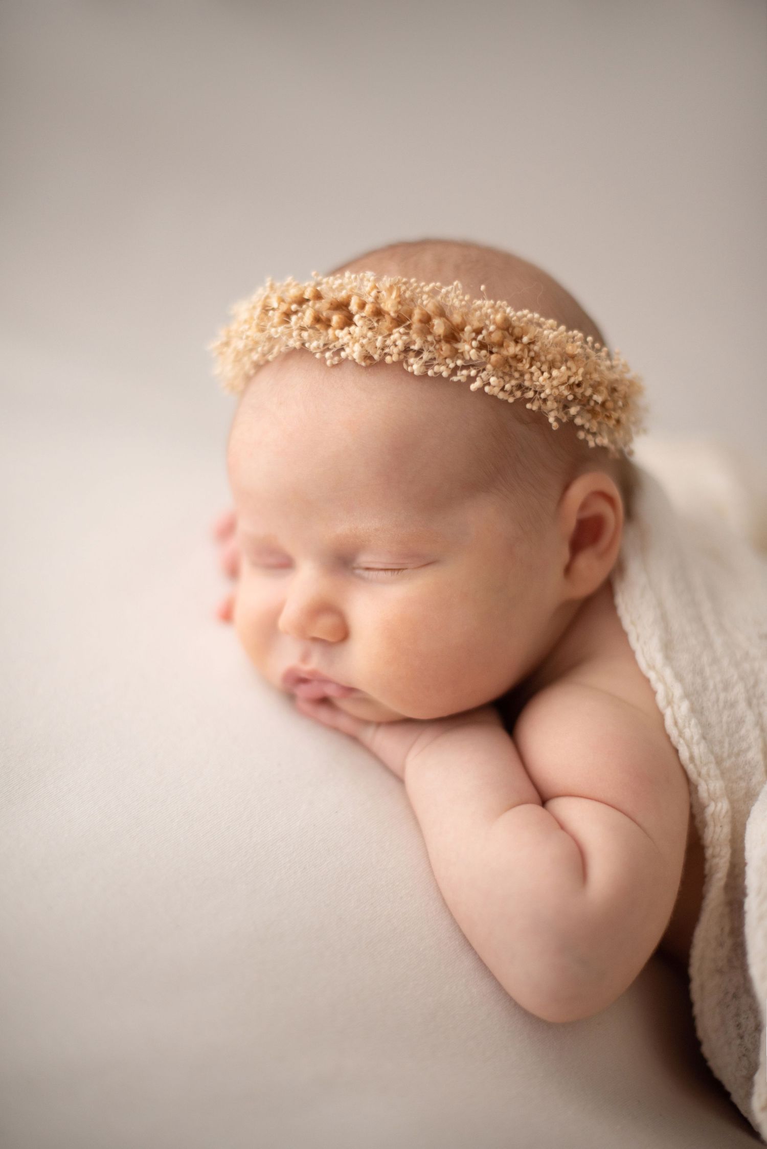 Sleeping newborn wearing beige floral crown lying peacefully on soft white backdrop during studio photography session.